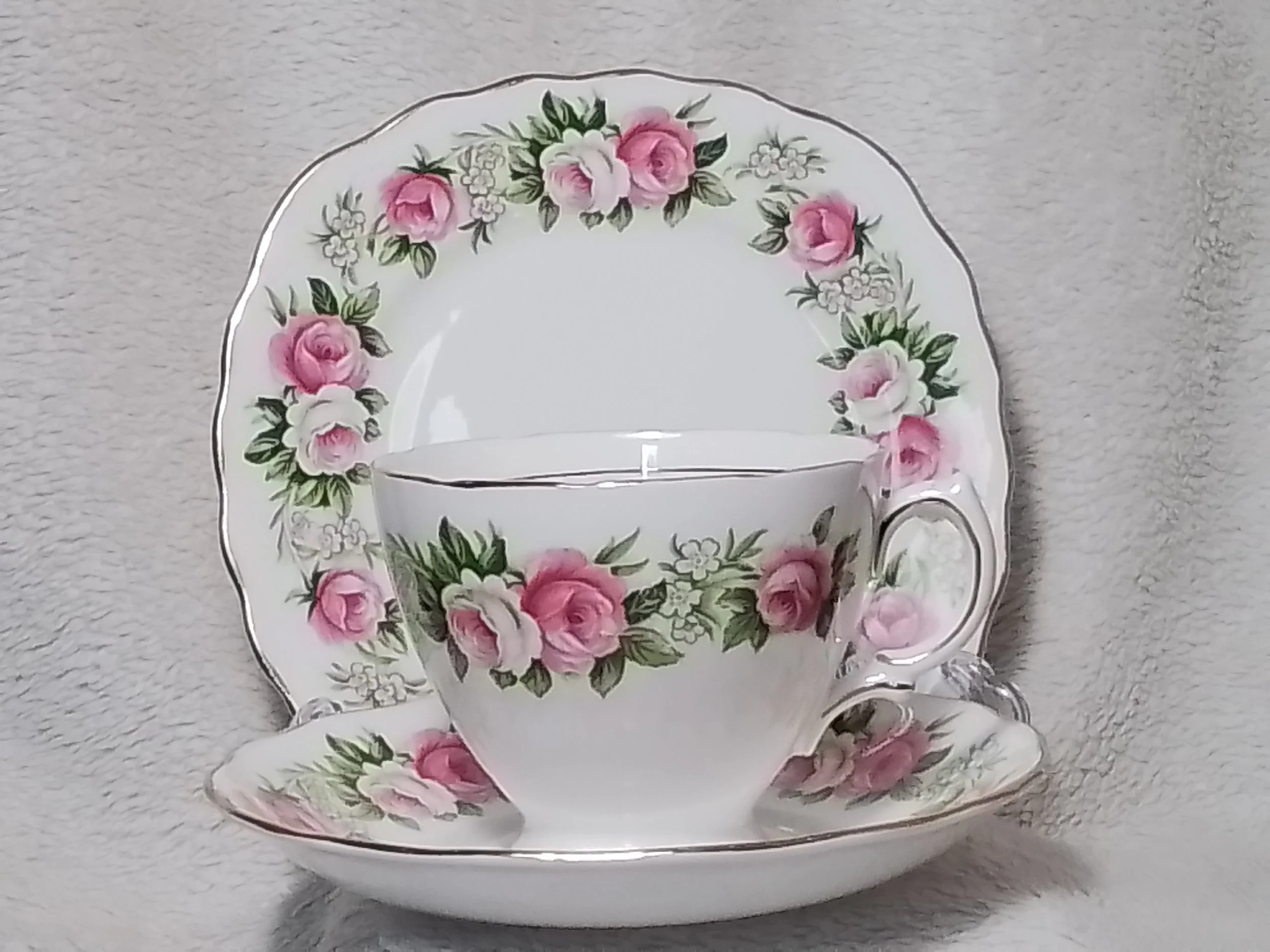 A floral patterned china teacup, saucer, and a dinner plate with pink roses and green leaves, set against a textured gray background.
