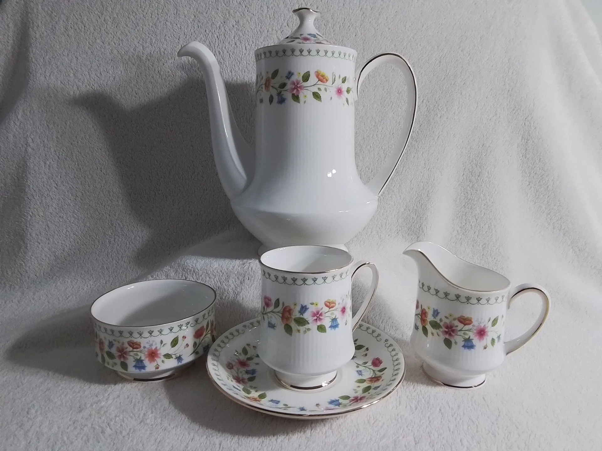 A porcelain tea set with floral patterns includes an urn-shaped teapot, a creamer, a cup with saucer, and a bowl, all with matching decorative borders.