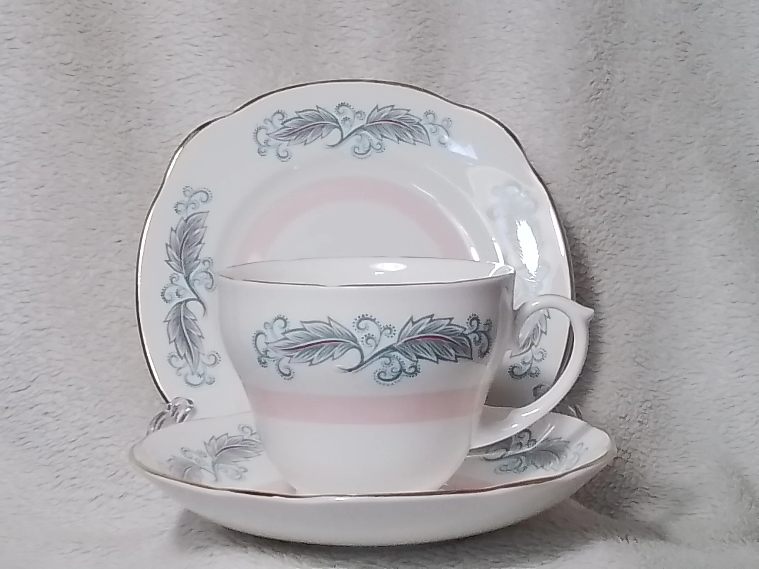 A white porcelain tea cup with floral patterns, placed on a matching saucer and behind a large matching plate, all with decorative gray and pink flowers, set against a textured gray background.