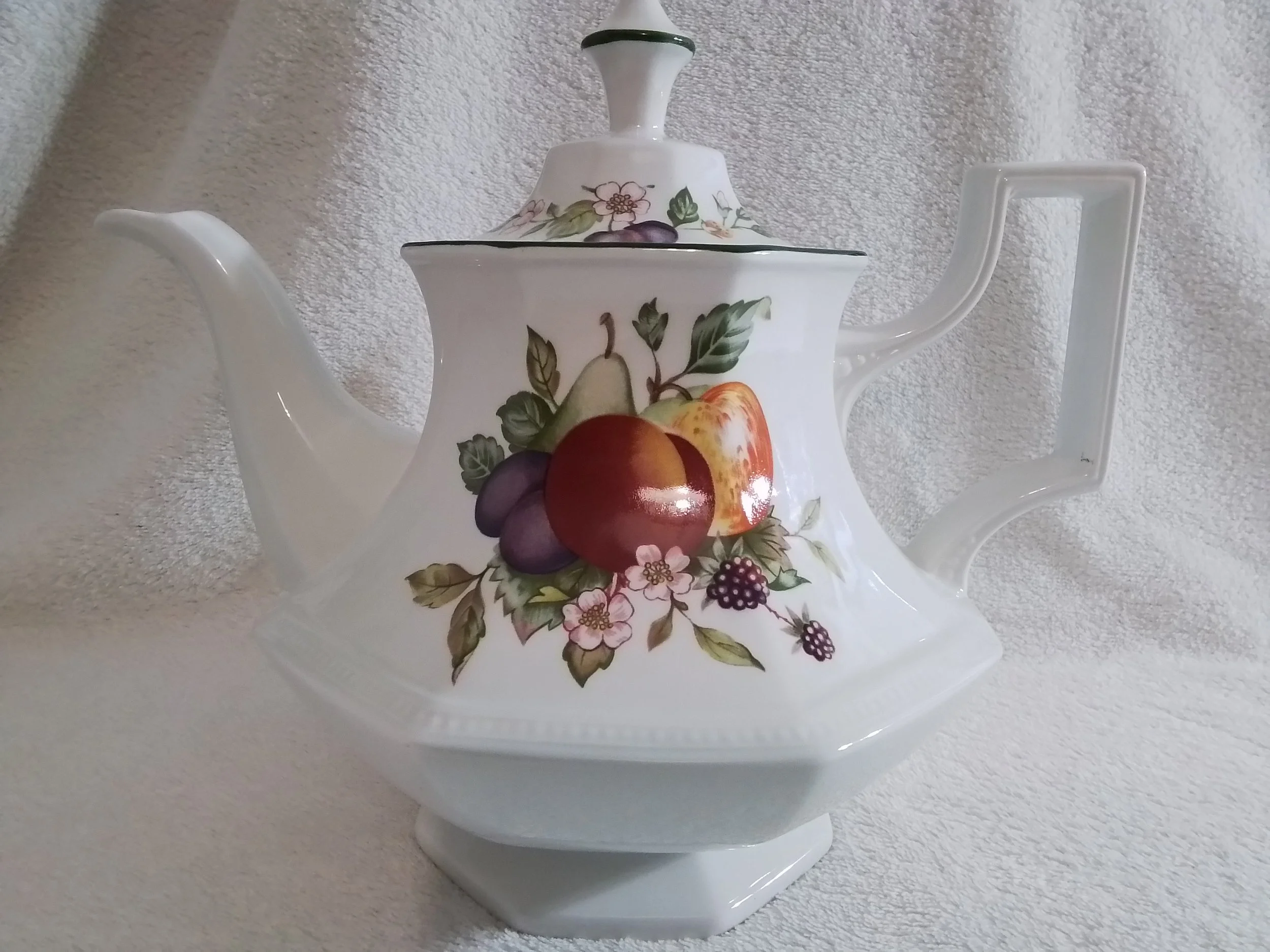 White ceramic teapot with a fruit and floral pattern, featuring images of an apple, pear, grapes, and a peach, set against a textured white background.