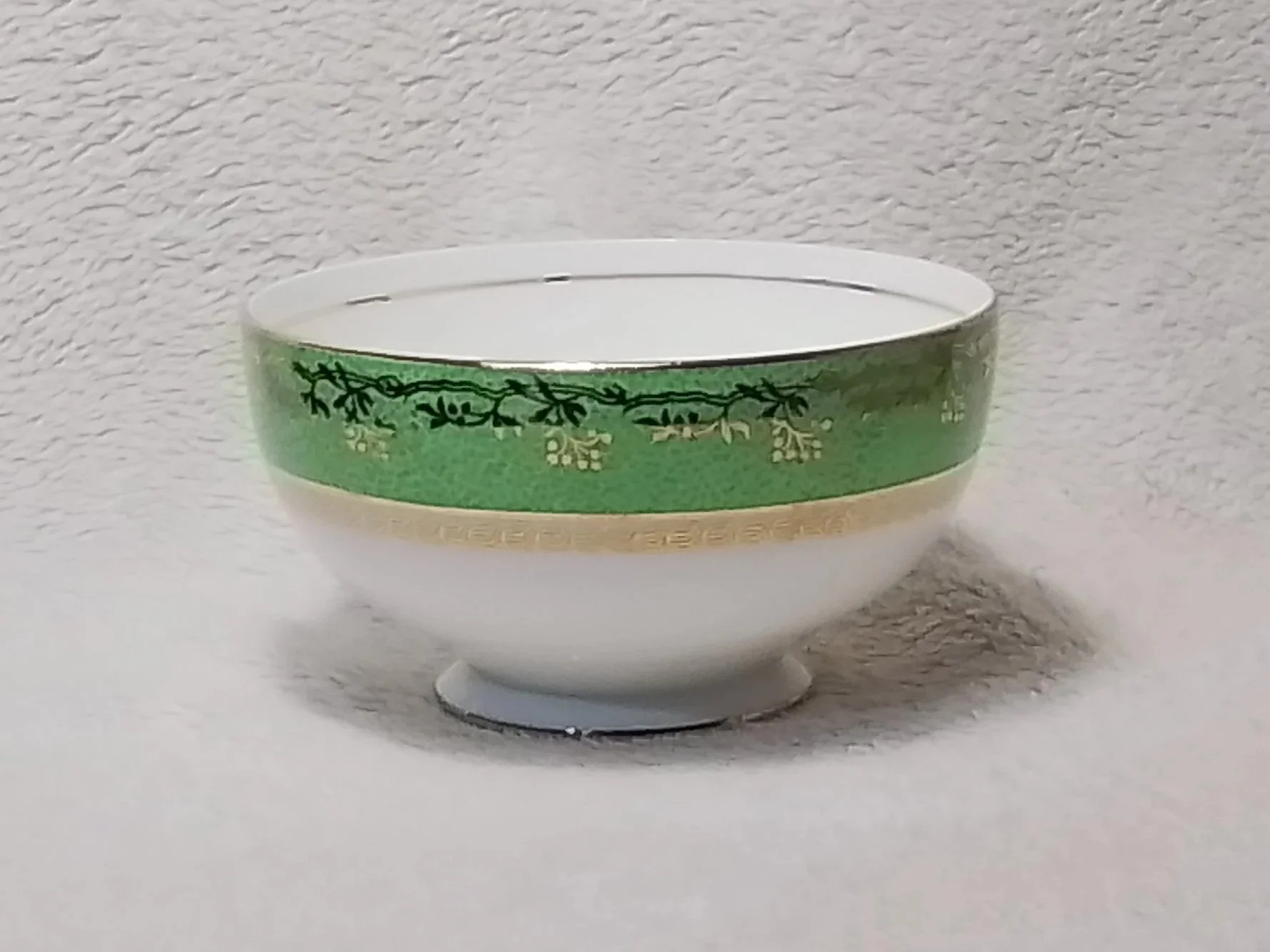 A small white porcelain bowl with a green decorative band and gold accents, placed against a plain textured background.