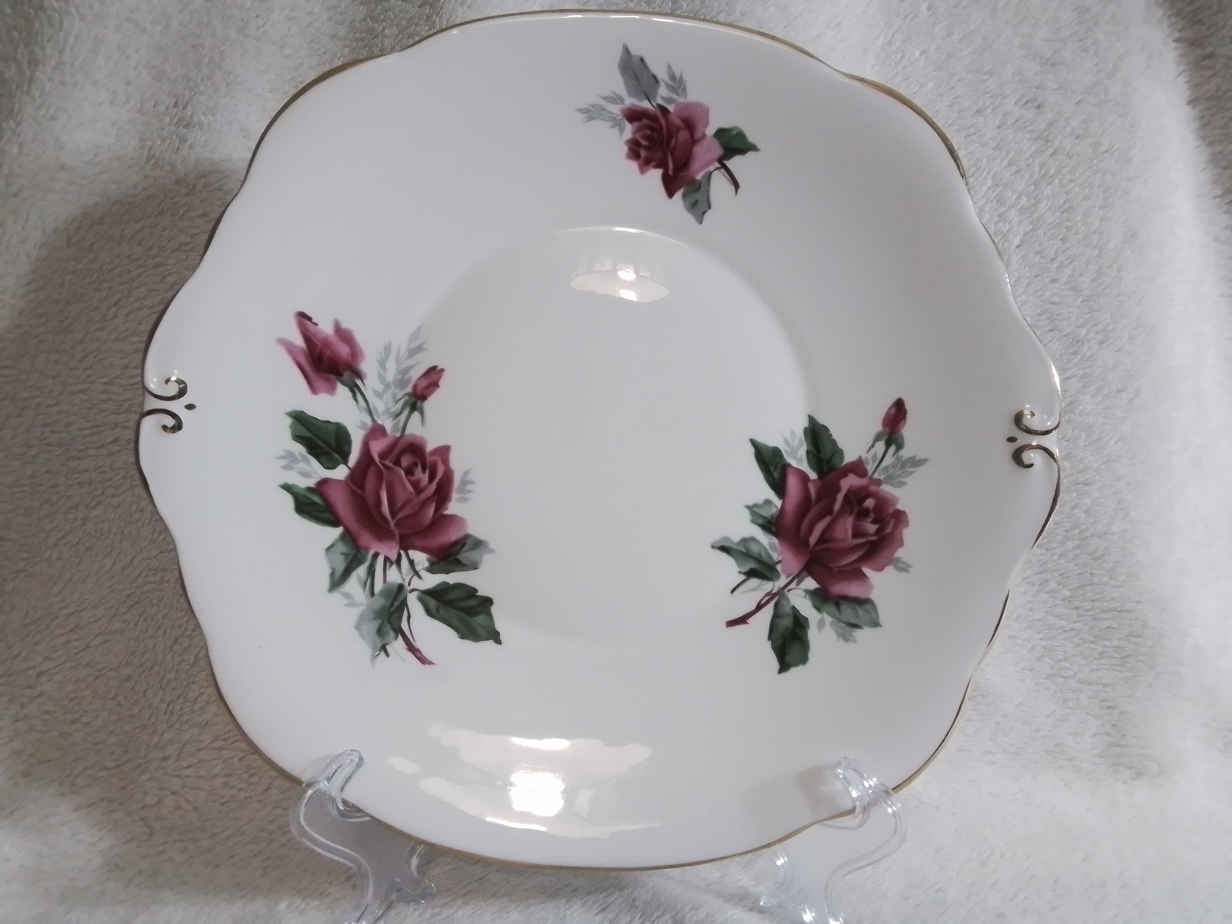 A white porcelain plate with a scalloped edge, decorated with pink roses and green leaves, with gold trim along the edge, displayed on a clear stand on a beige textured surface.