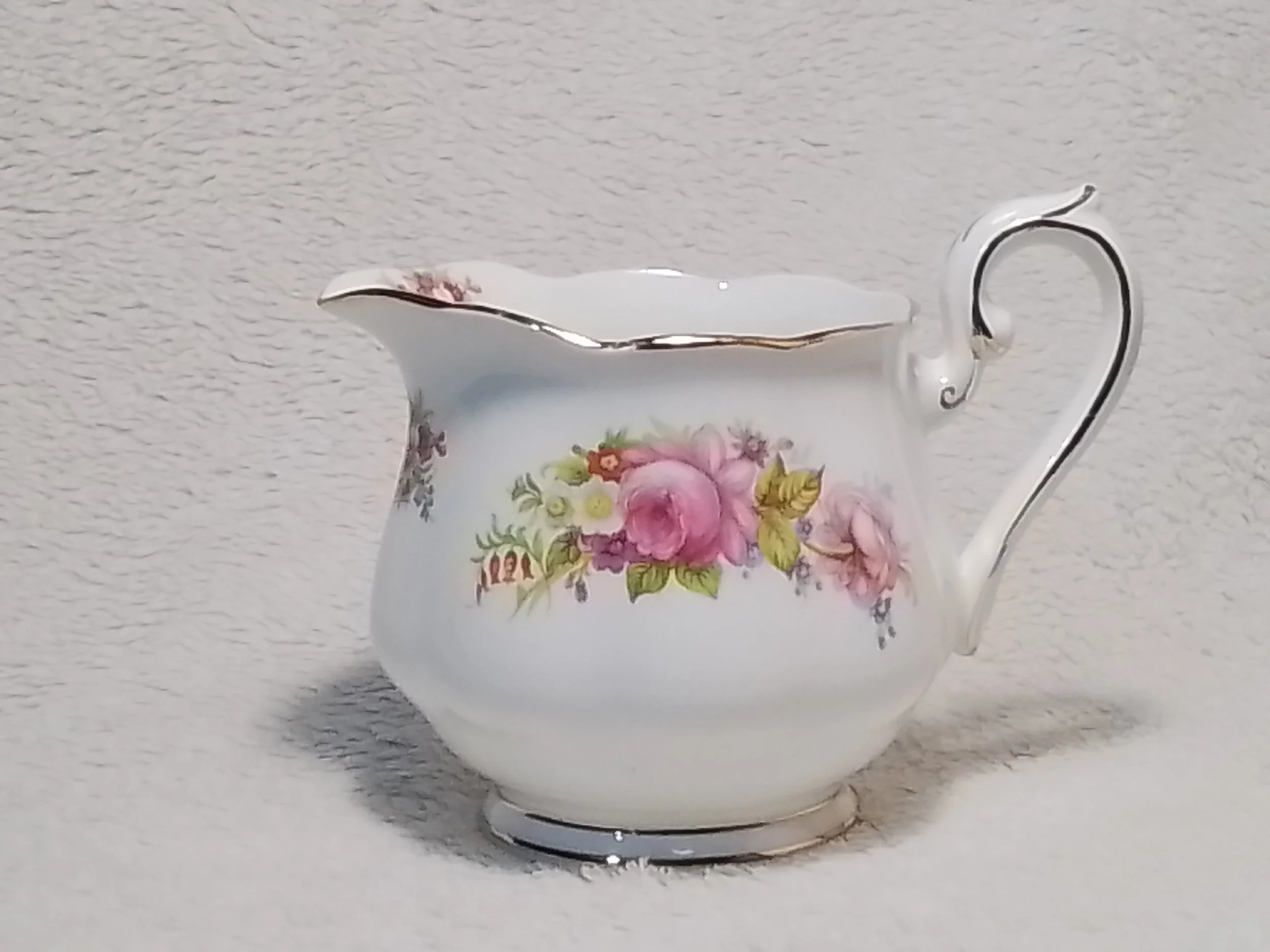 A decorative white porcelain gravy boat with floral designs, gold trim, and an ornate handle, placed on a textured white surface.