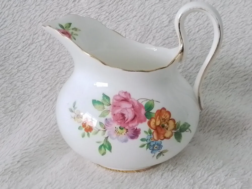 White porcelain creamer with pink, orange, blue, and green floral pattern, gold trim, and a curved handle, placed on a soft, textured surface.