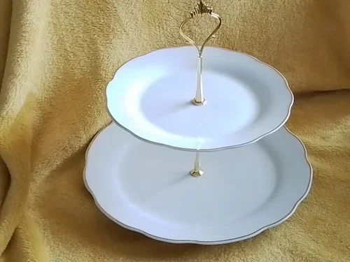Two-tier white ceramic serving stand with a silver handle on a gold fabric background.