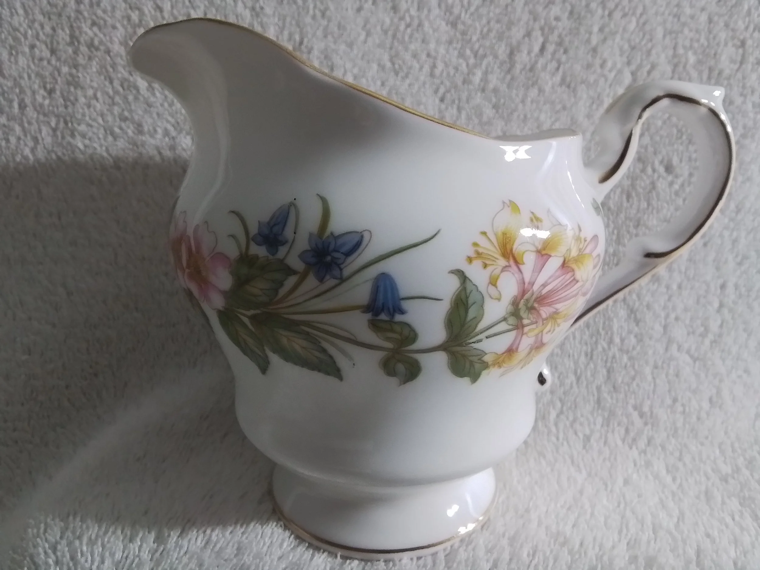 A white ceramic gravy boat decorated with pink, blue, and yellow floral patterns, placed on a textured white surface.