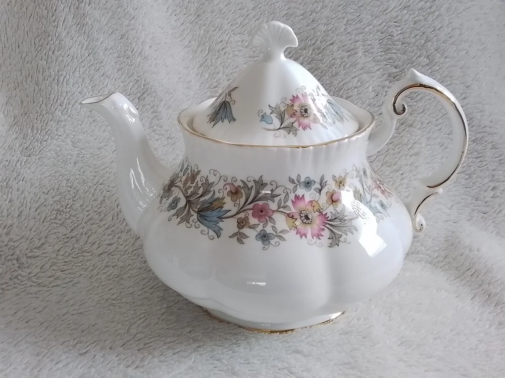 A white porcelain teapot with floral patterns and gold trim rests on a soft, light-colored surface.