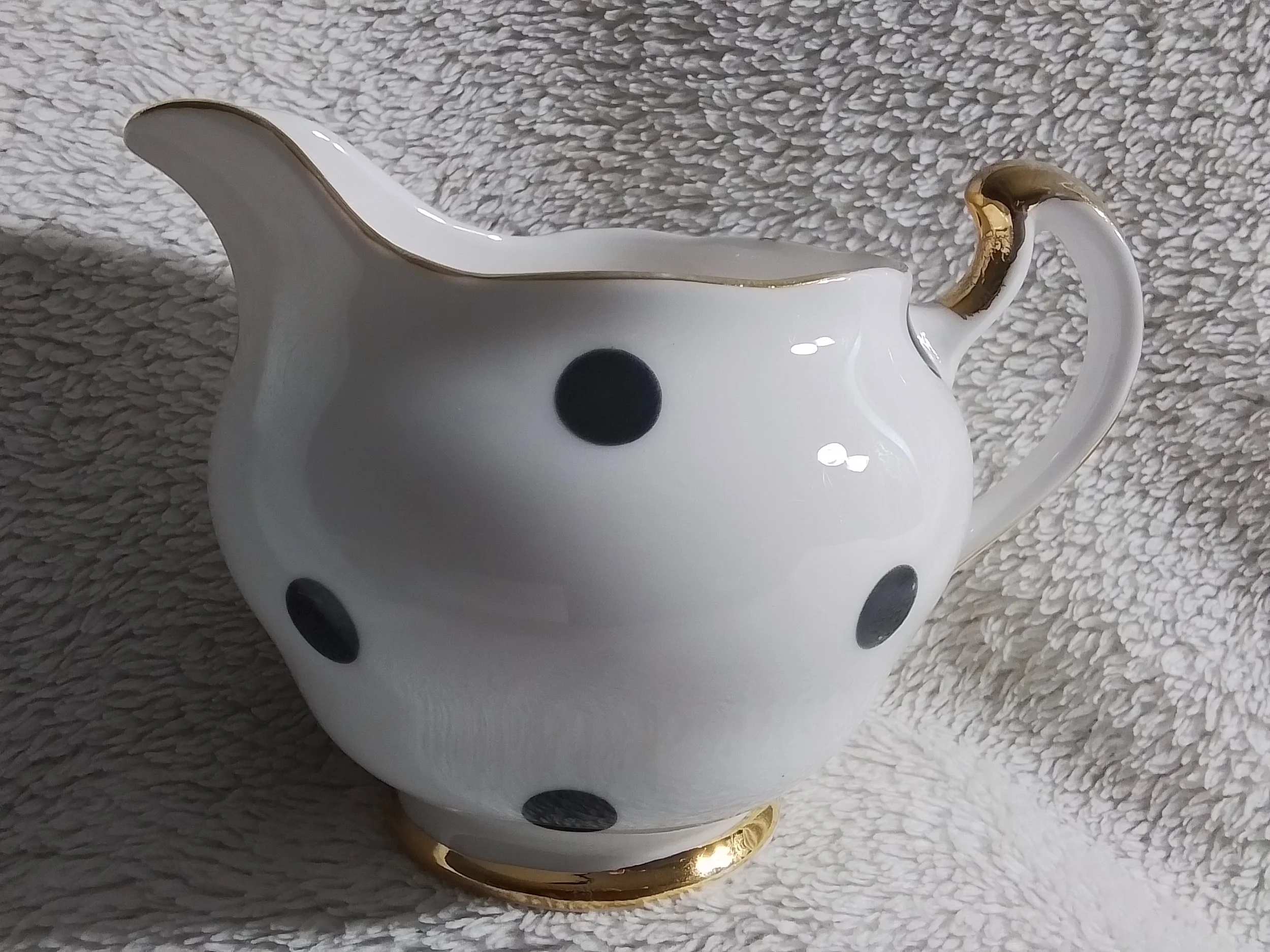 A white ceramic teapot with black polka dots and gold accents on the handle and base, placed on a textured fabric surface.