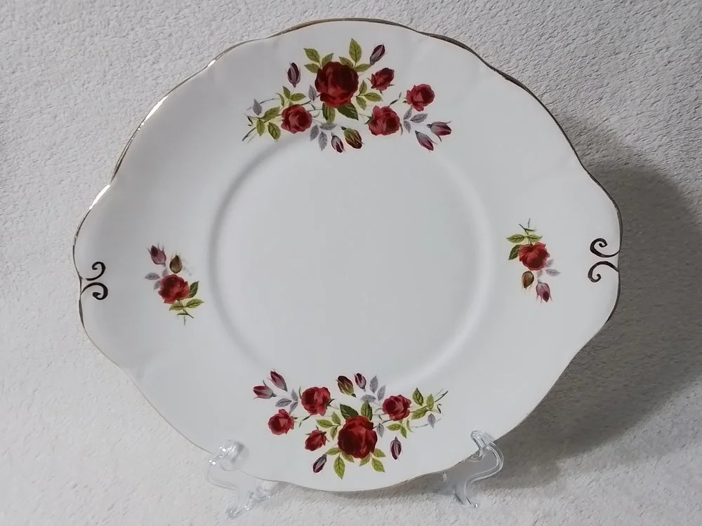 A white decorative plate with floral patterns of red roses and green leaves, displayed on a clear stand against a light-colored background.