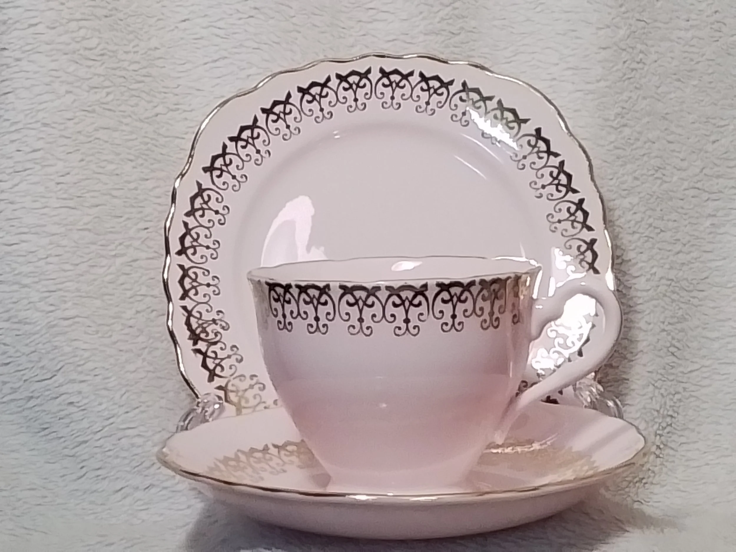 A white porcelain teacup with a gold decorative pattern around the rim, placed on a matching saucer with the same pattern, and a larger matching plate behind them, set against a textured light gray background.