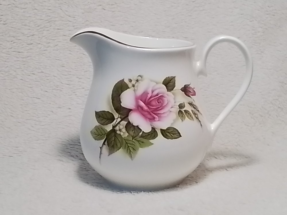 White ceramic pitcher with a pink rose and green leaves design, set against a plain light background.