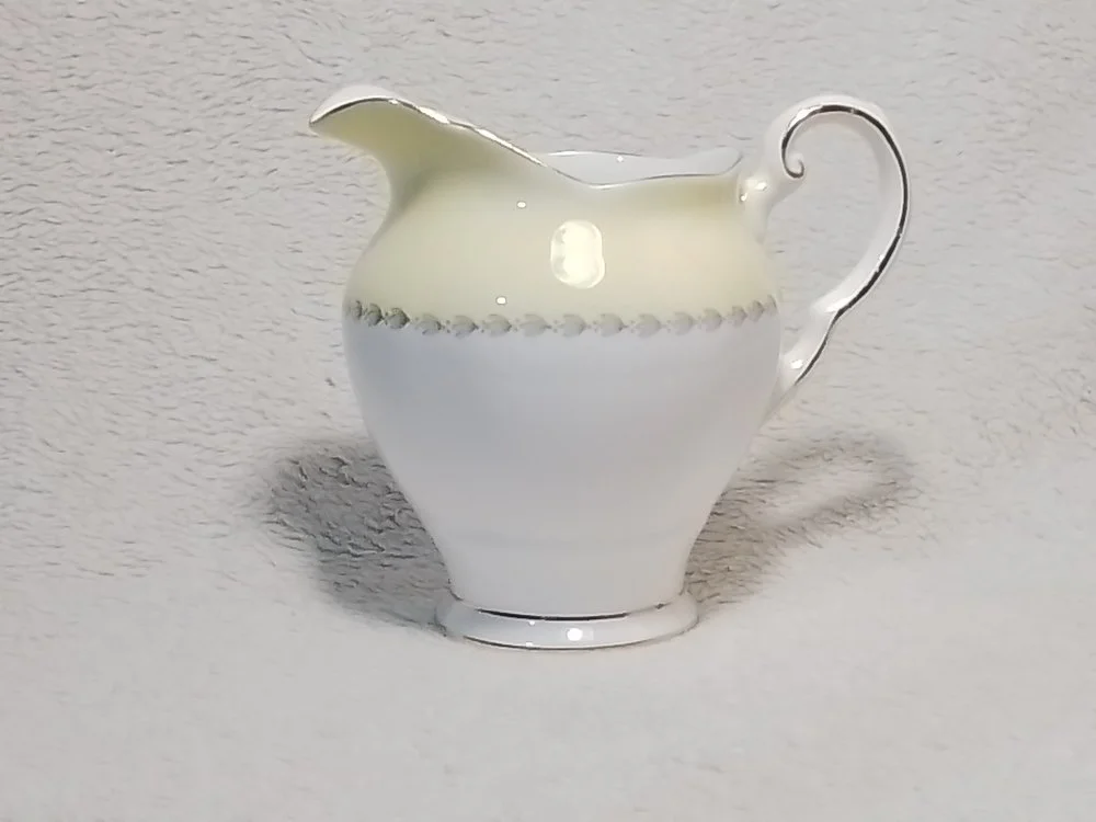 A white porcelain creamer with a decorative silver band near the top, placed on a light-colored surface.