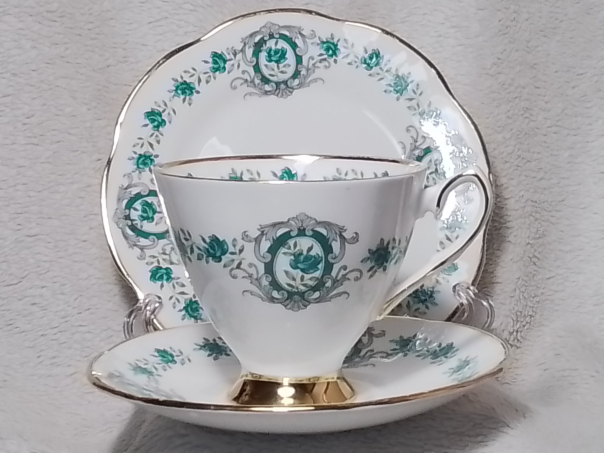 A fine porcelain tea cup, saucer, and plate set with a ornate teal and gray floral pattern and gold accents, displayed against a light gray background.
