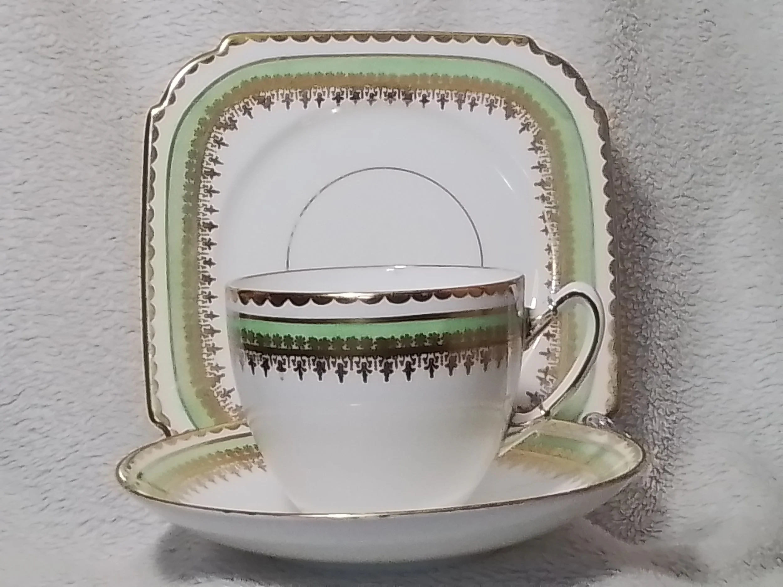 A fine china tea cup and matching saucer, both decorated with gold, green, and black patterns, placed in front of a matching rectangular and an oval plate with similar decorative patterns.