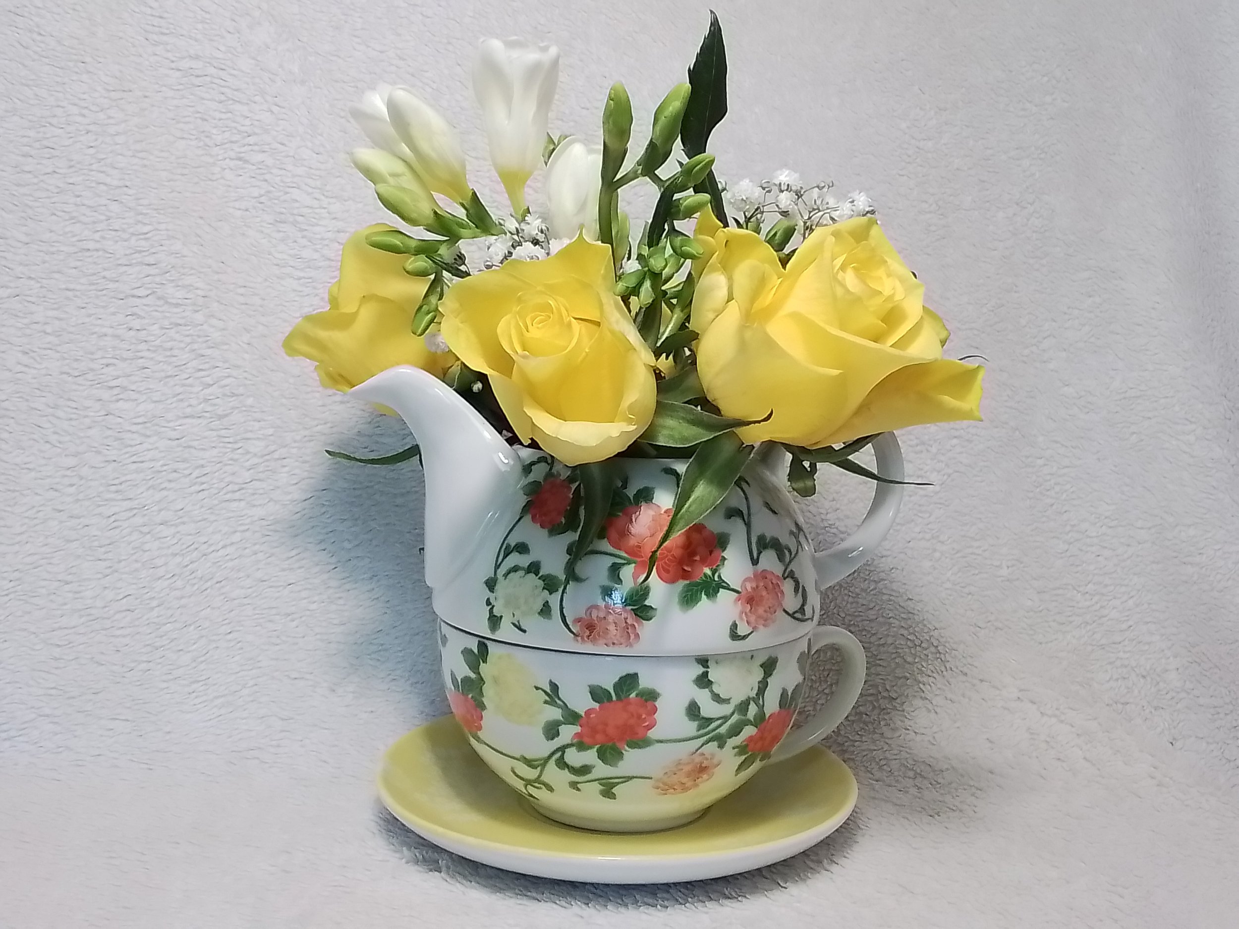 A floral arrangement with yellow roses, white flowers, and green leaves in a teapot-shaped vase with a floral pattern, placed on a small yellow tray against a light-colored textured background.
