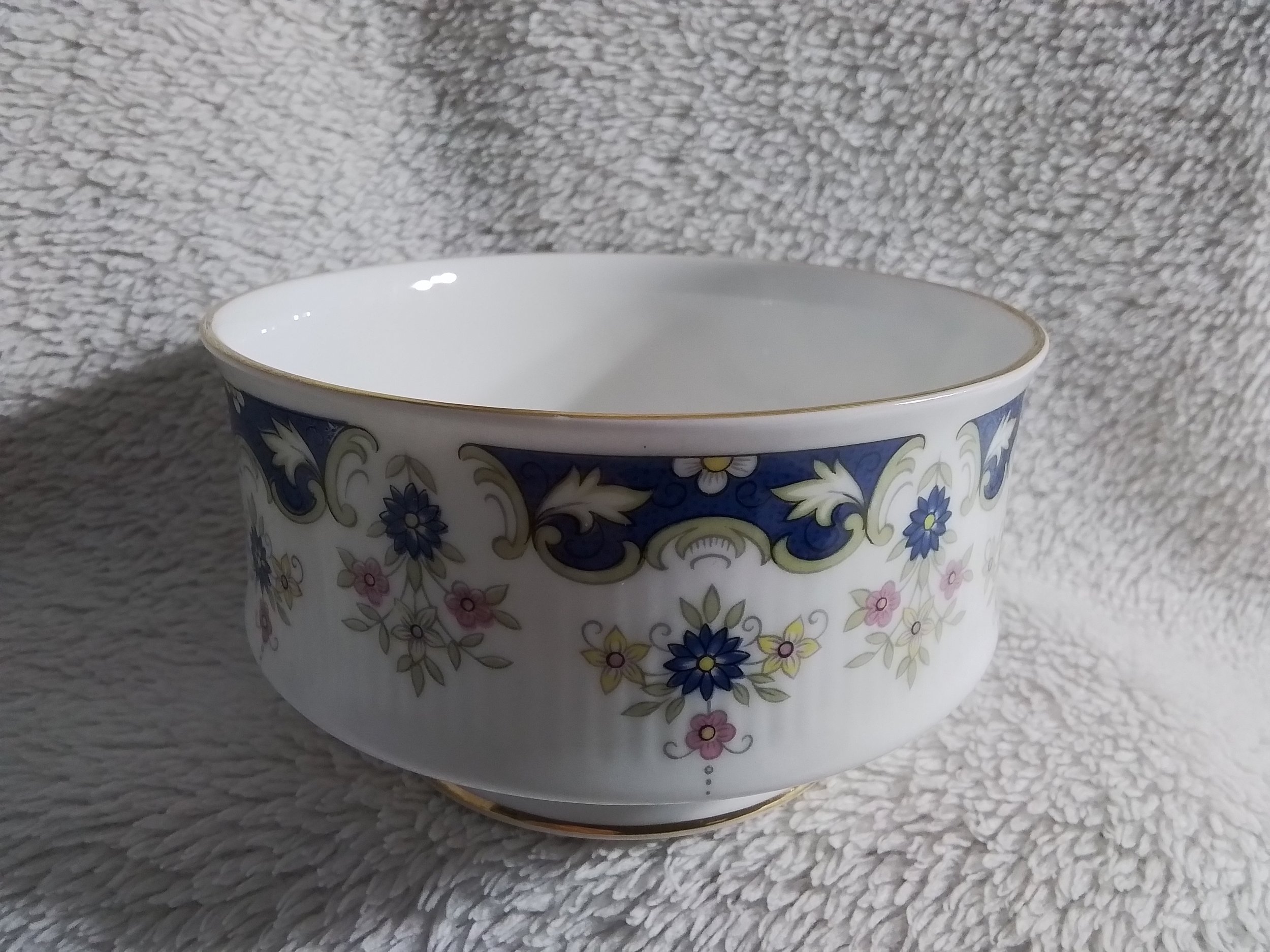 A decorative porcelain bowl with floral and scroll patterns in blue, yellow, pink, and gold on a white background, resting on a textured beige surface.