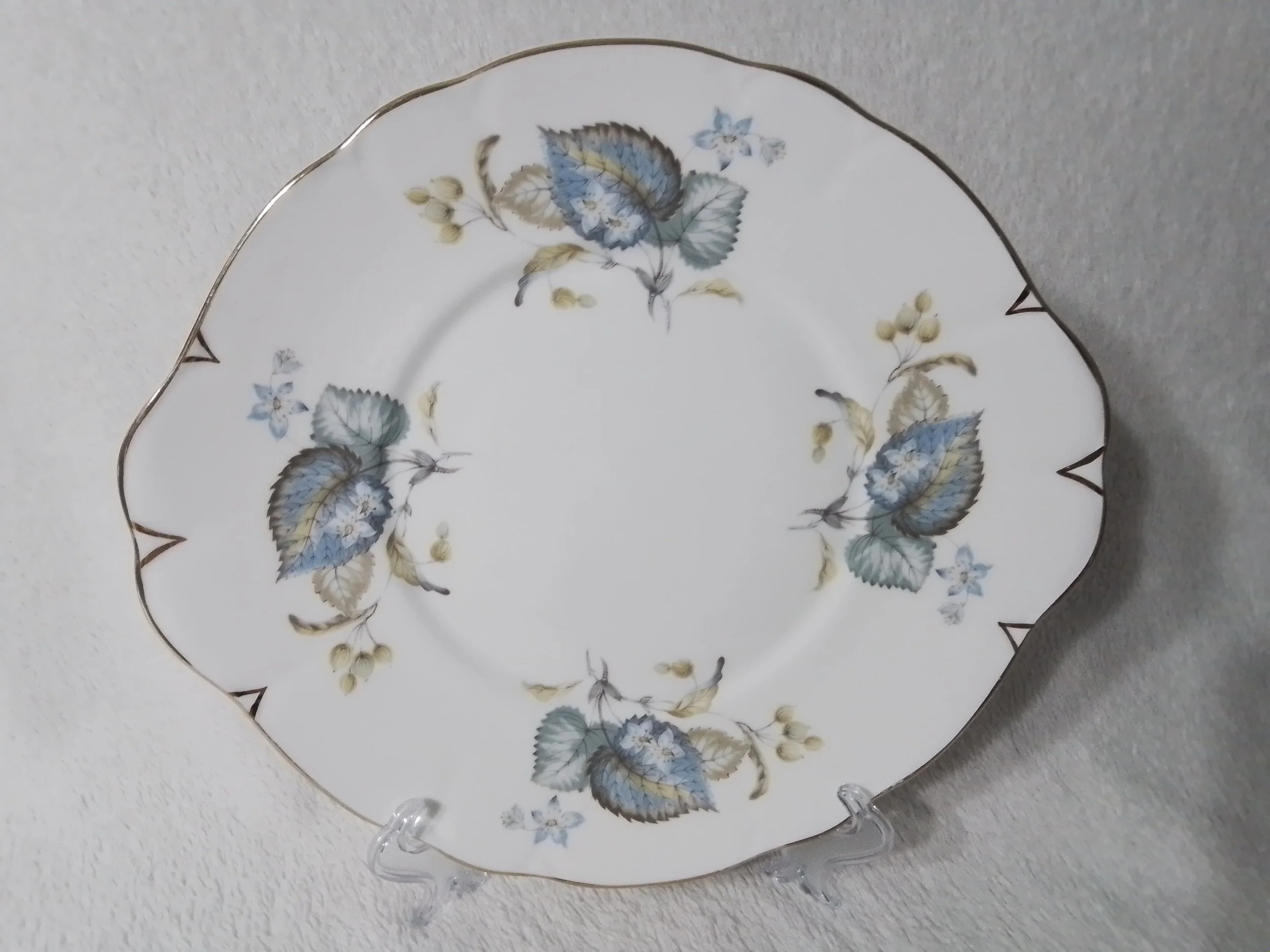 Decorative porcelain plate with blue and beige floral pattern, gold trim, and a scalloped edge displayed on a clear plastic stand.