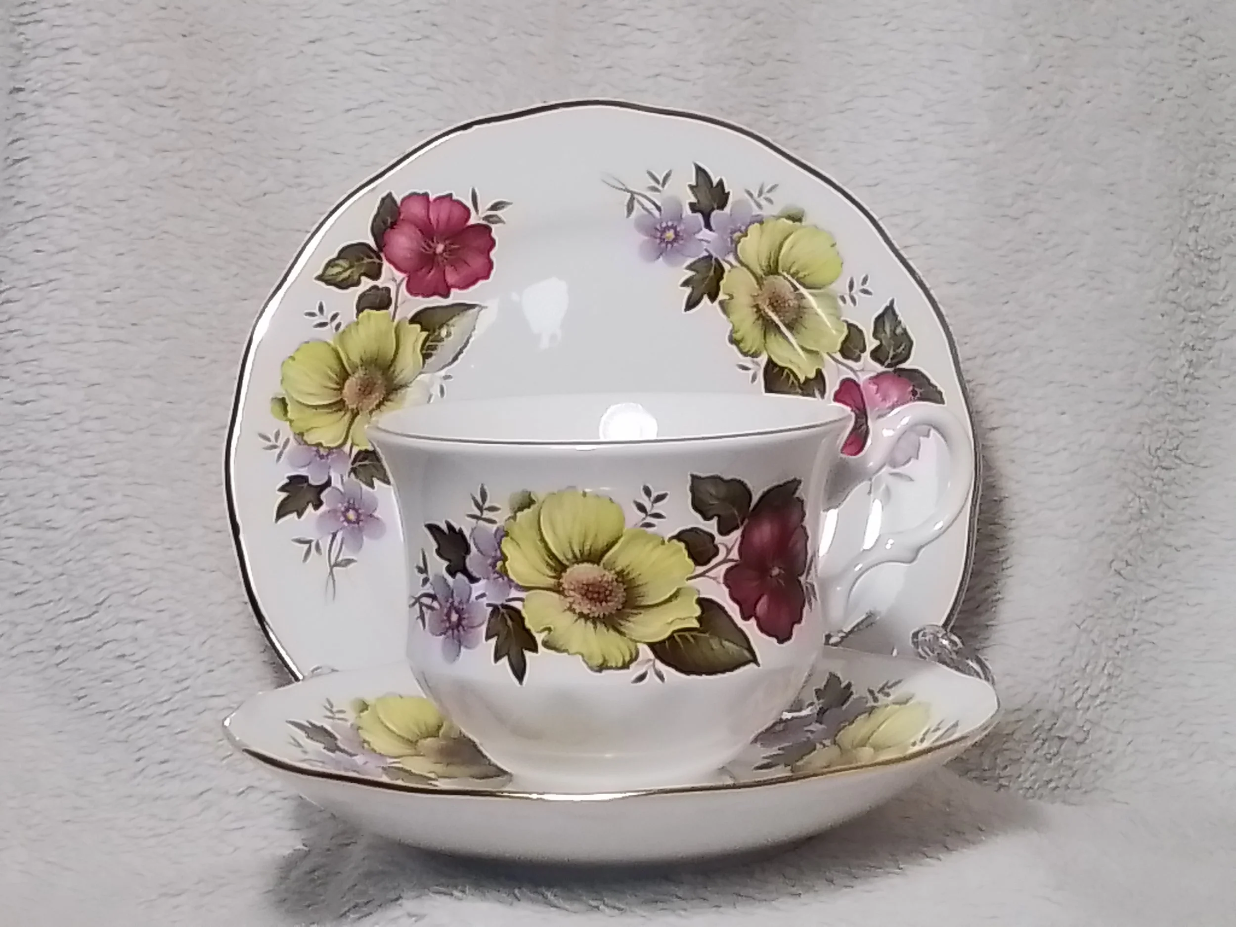 A floral patterned tea cup and saucer set with flowers in shades of yellow, pink, and purple, placed on a textured white surface.