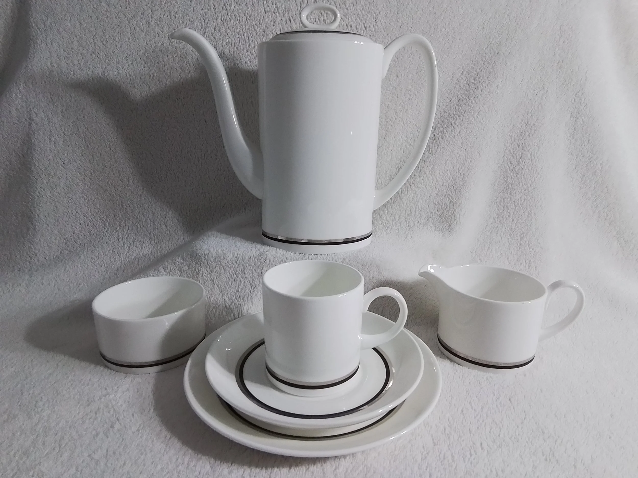 A white teapot, teacup, bowl, creamer, and plates arranged on a white textured surface.