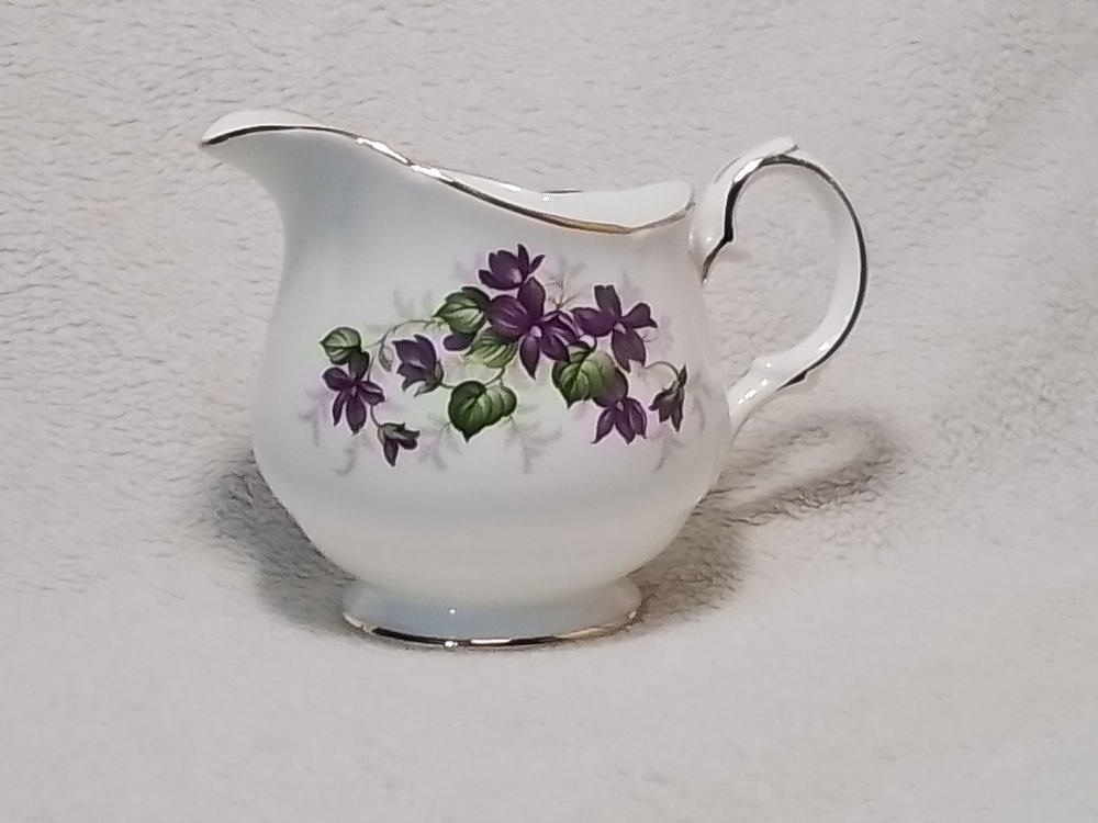 A white ceramic creamer pitcher with purple floral design and gold trim, placed on a soft light-colored fabric background.