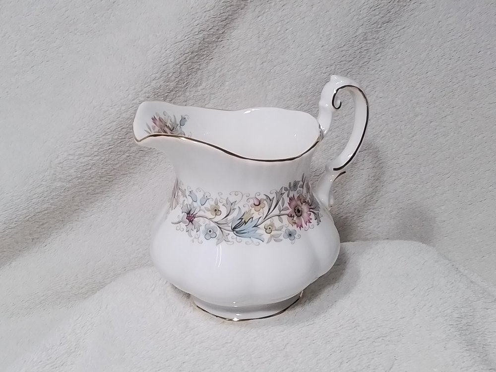 White porcelain creamer with floral pattern and gold accents on a soft, light-colored fabric background.