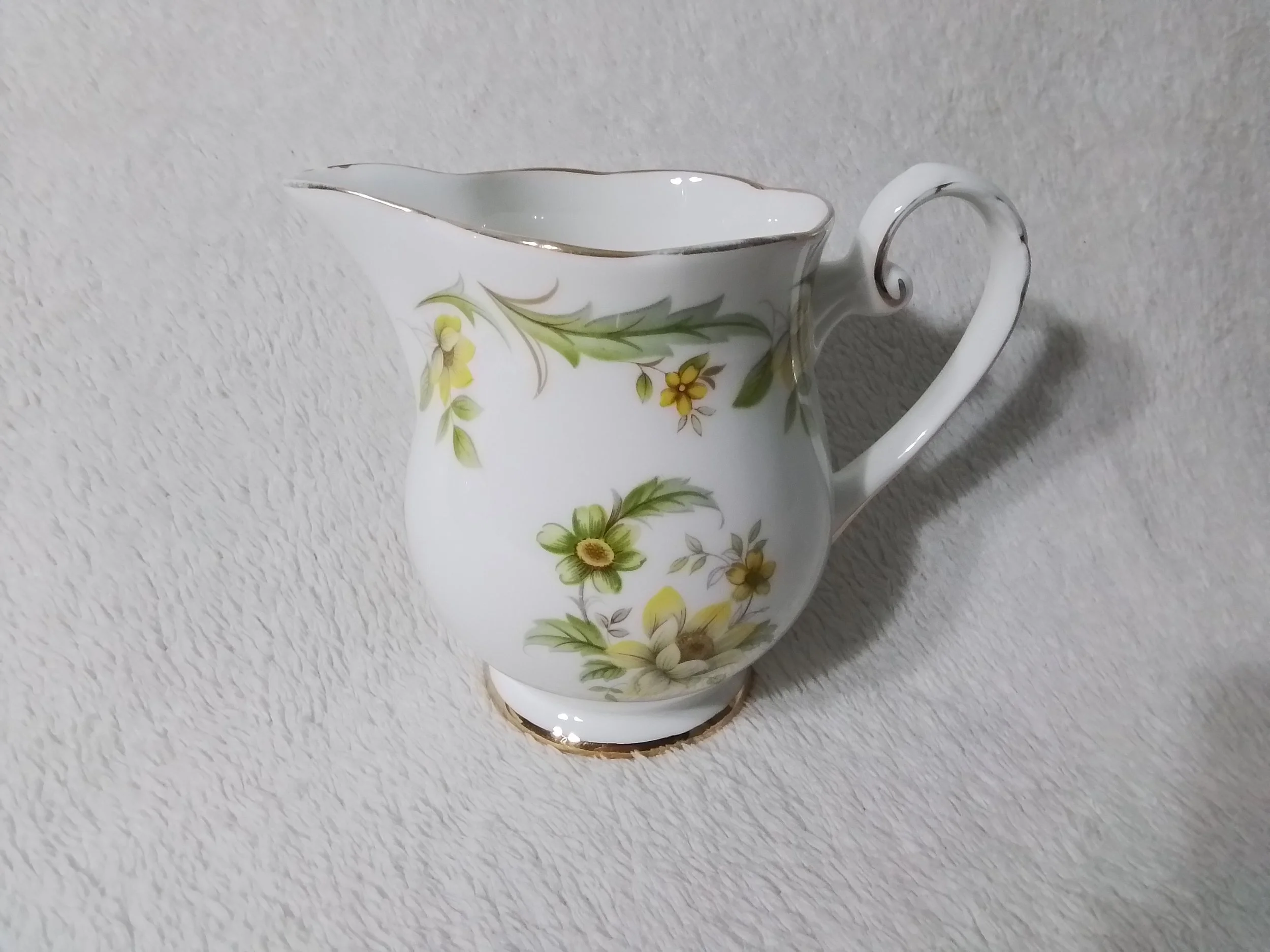 A white ceramic pitcher with a floral design of yellow and green flowers and leaves, and gold accents along the rim, handle, and base, placed on a textured white surface.