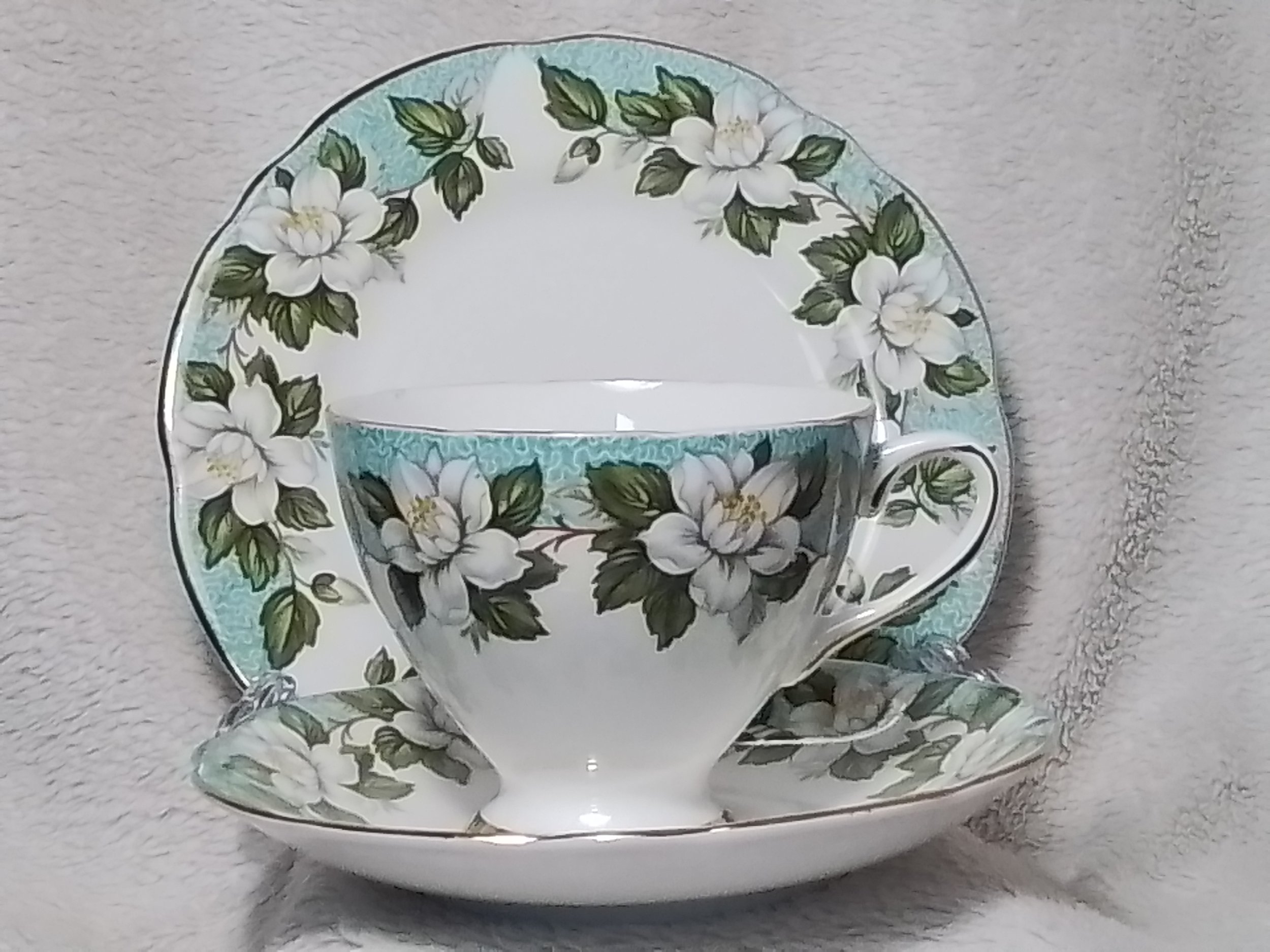 A teacup with a floral pattern of white flowers and green leaves, placed on a matching saucer against a beige fabric background.