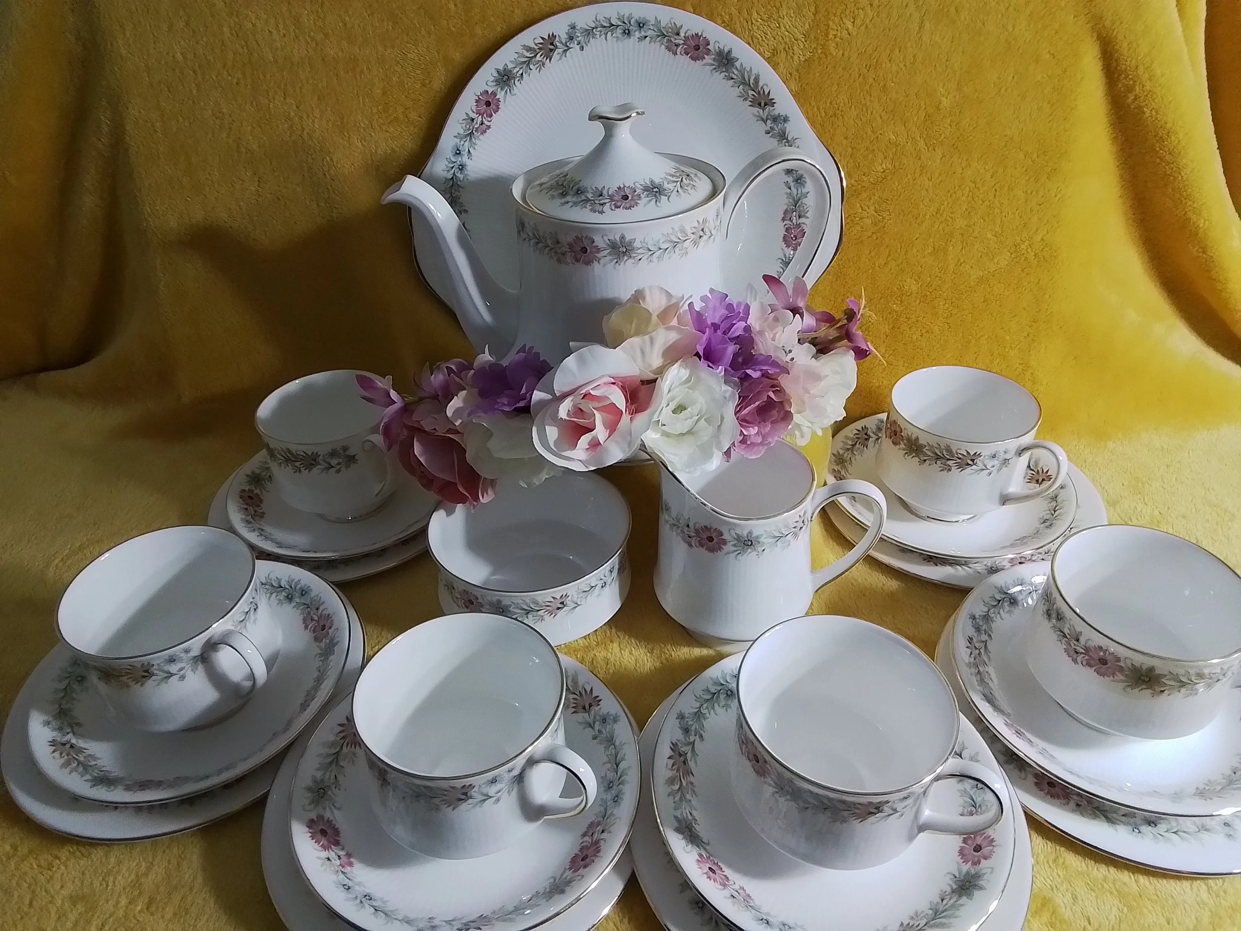 A vintage tea set with cups, saucers, a teapot, a creamer, and a tray, all decorated with a floral pattern, arranged on a yellow cloth with a bouquet of pink and white flowers in the center.