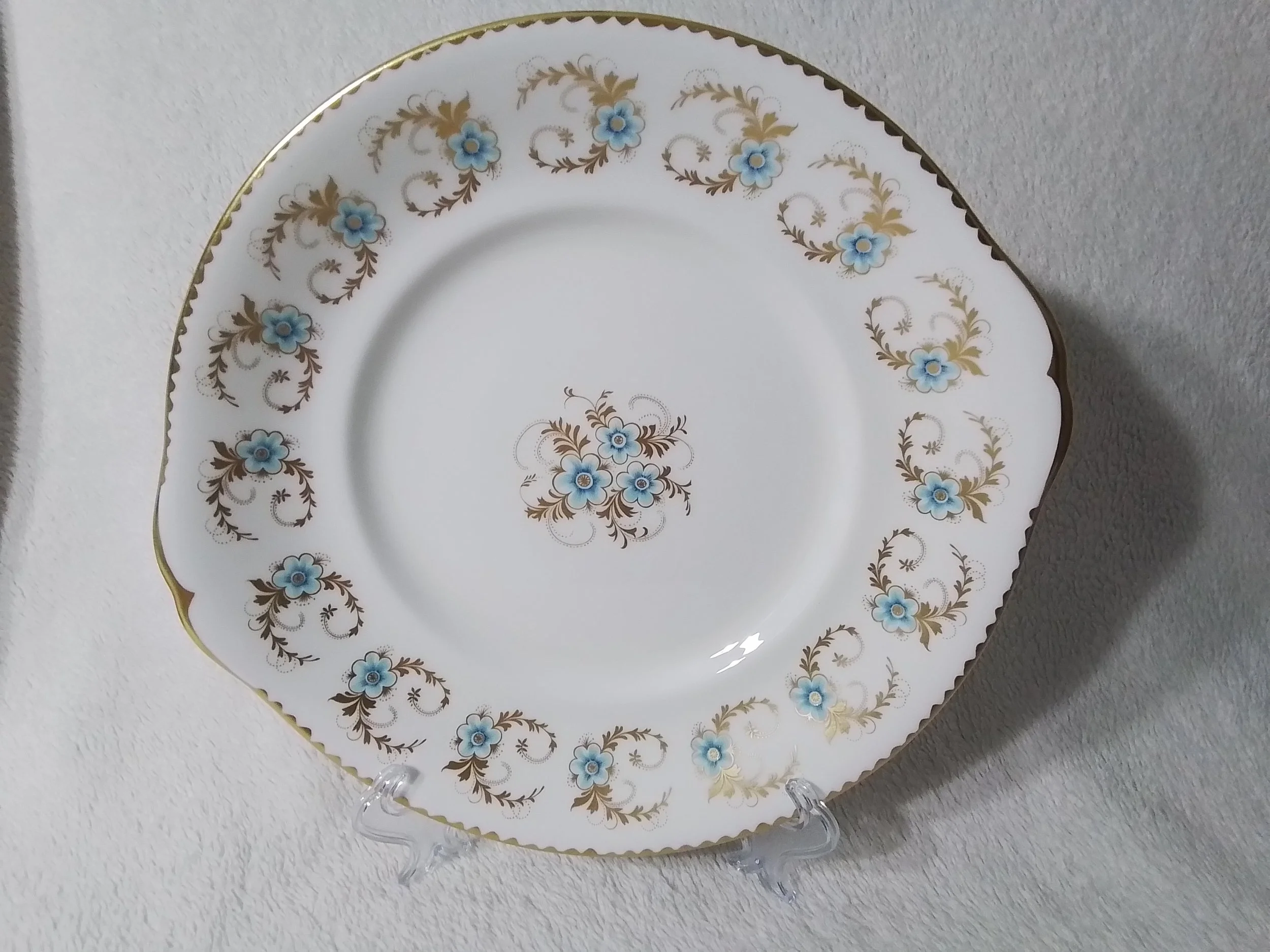 A decorative porcelain serving plate with blue floral patterns and gold accents, displayed on a clear plastic stand against a white textured background.