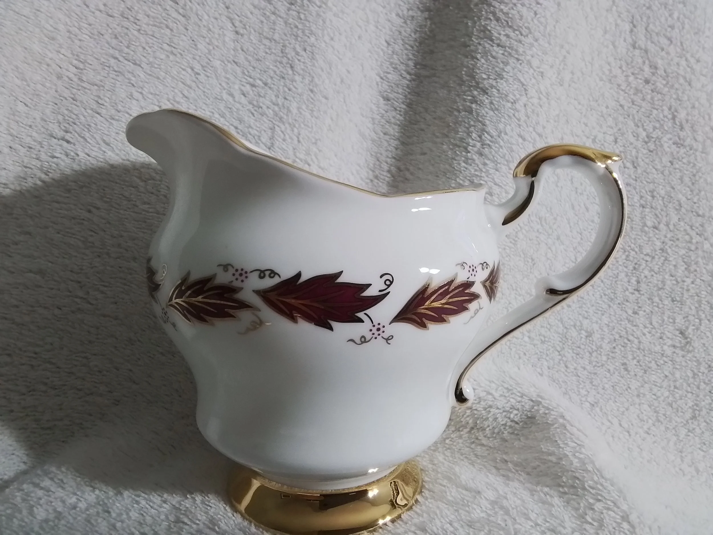 A white porcelain gravy boat with a gold rim and a floral leaf pattern in shades of burgundy and gold, sitting on a white textured surface.