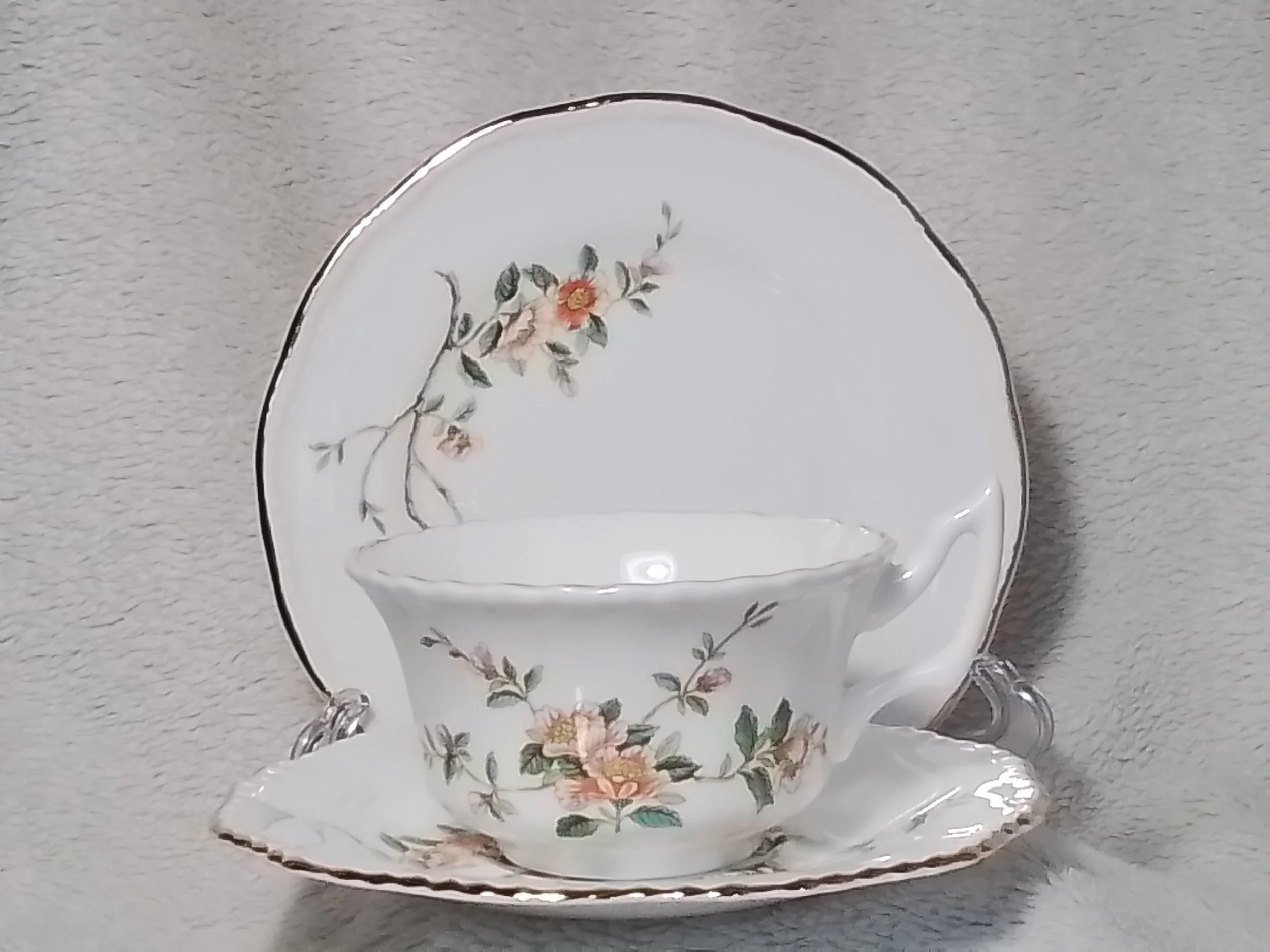 A vintage floral tea cup and saucer set with a matching dessert plate, decorated with pink and peach flowers and green leaves, displayed against a light fabric background.