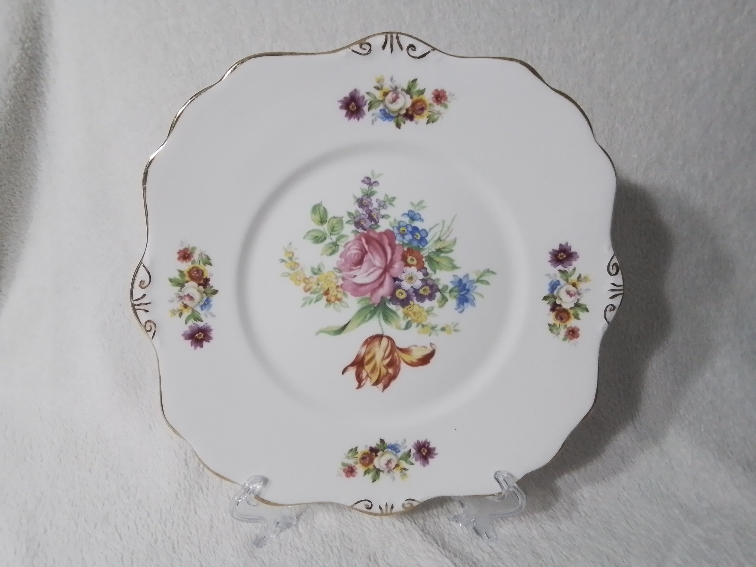 Decorative porcelain plate with floral patterns, gold trim, and a central bouquet of pink, purple, yellow, and blue flowers displayed on a clear stand against a light-colored wall.