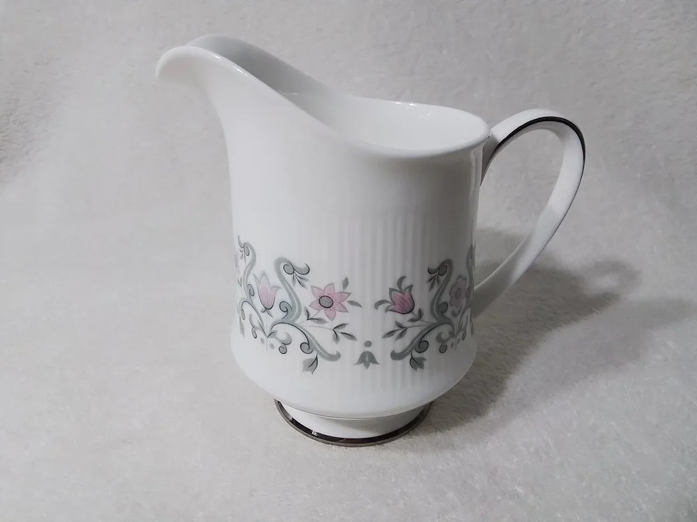 White ceramic creamer with pink and gray floral pattern, handle on right side, on a light textured surface.