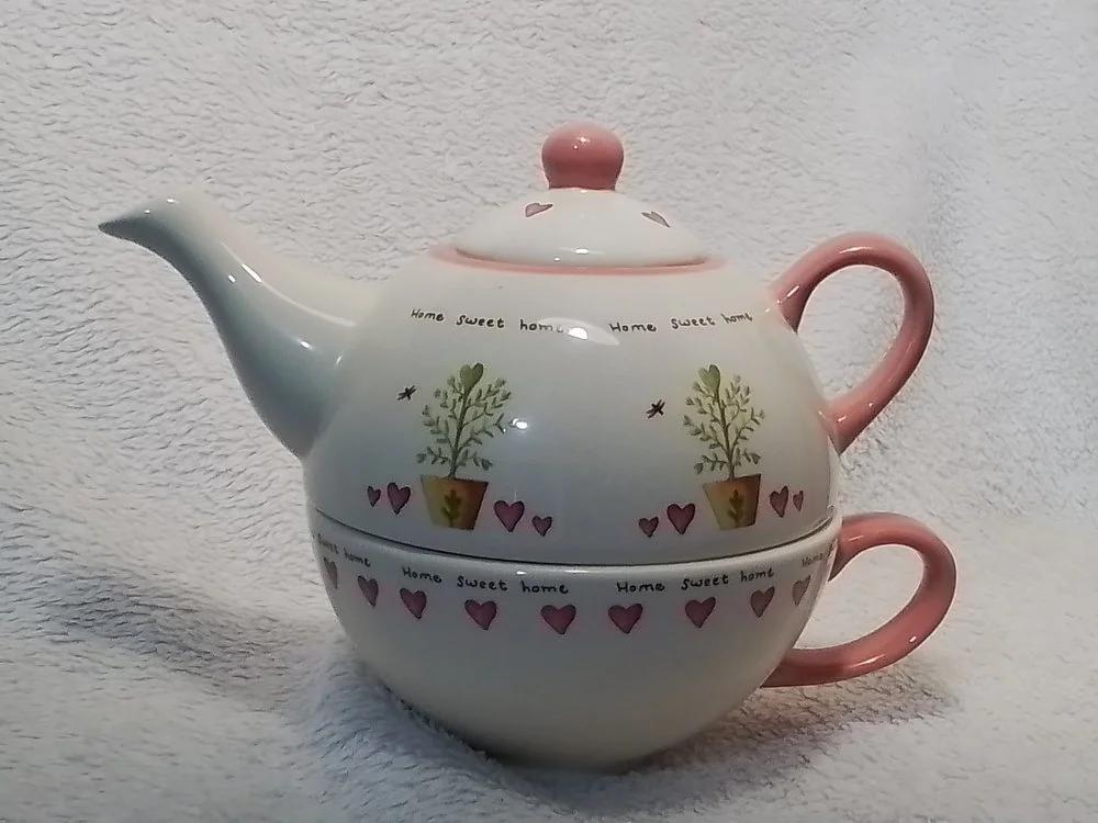 A teapot with a pink handle and lid, decorated with small trees in pots, hearts, and the words 'Home sweet home' on a white textured background.