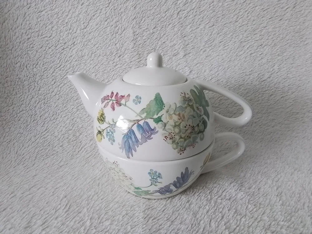 A white ceramic teapot and creamer stacked, with floral and leaves pattern, on a textured light gray surface.