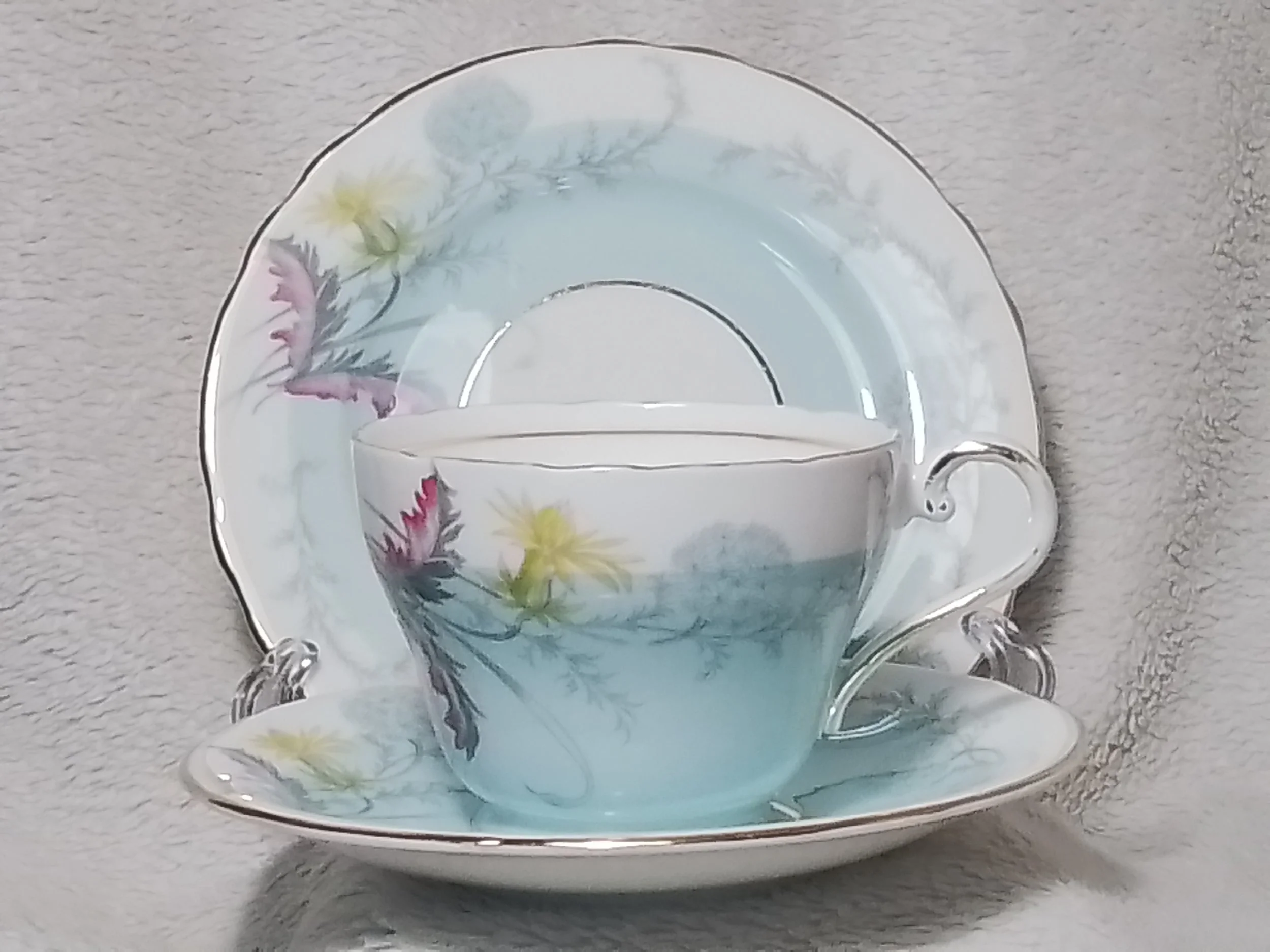 A porcelain tea set consisting of a teacup with a matching saucer and a large plate, all decorated with a floral pattern featuring pink and yellow flowers, set against a soft, neutral background.