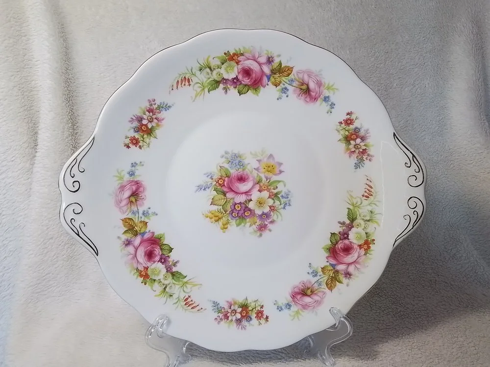 A decorative porcelain plate with a floral pattern, featuring pink and yellow roses and smaller flowers, with black swirl accents on the scalloped edges, displayed on a clear stand.