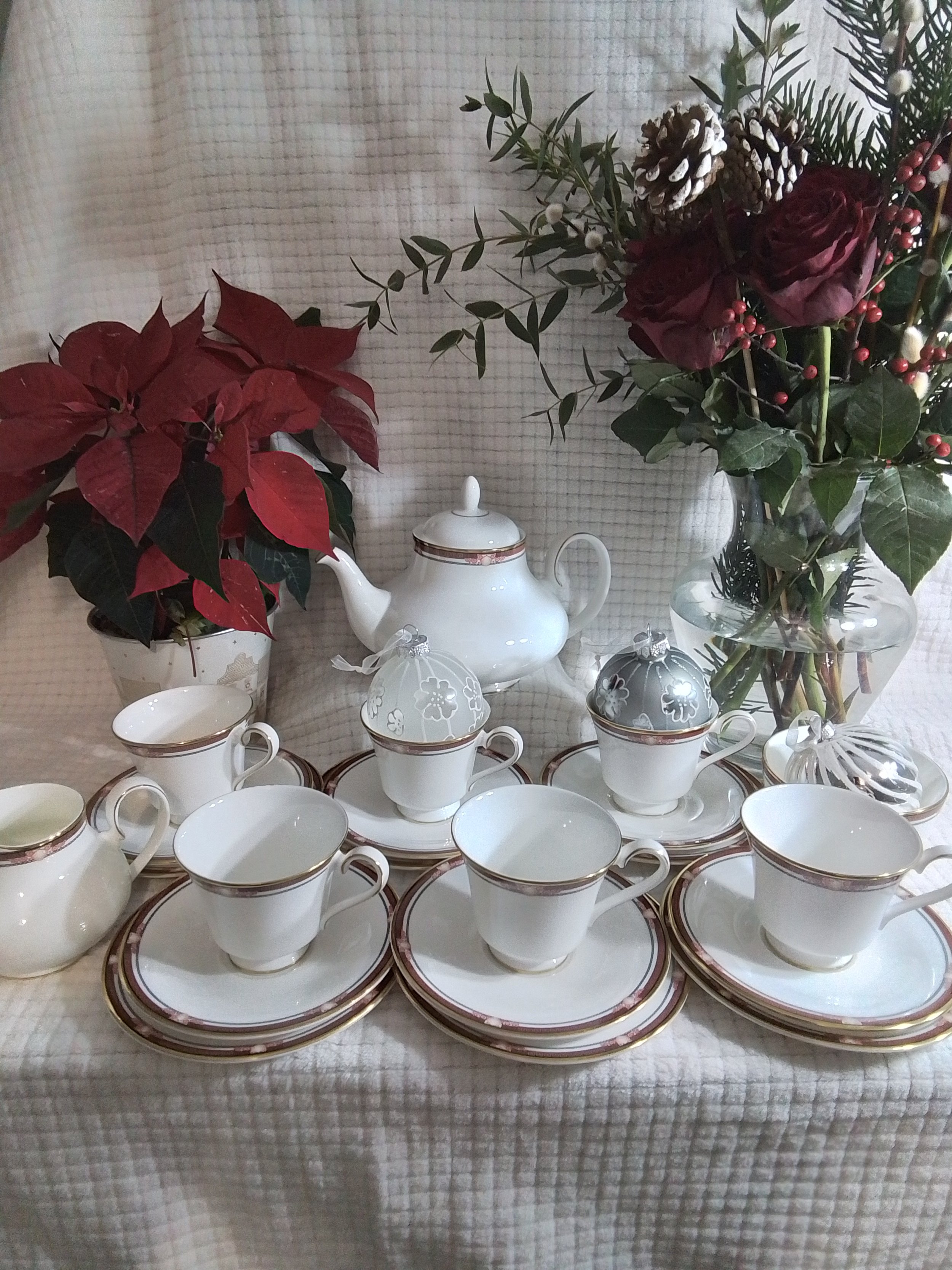 A festive table setting with white china teacups, saucers, and a teapot, all with a gold and pink trim, arranged on a beige tablecloth. There are two vases with poinsettia and other greenery decorations in the background, along with pinecones and berries.