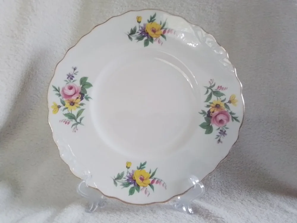 A white porcelain plate with a floral pattern featuring pink, yellow, and purple flowers and green leaves, with a scalloped gold rim, displayed on a clear stand against a light background.