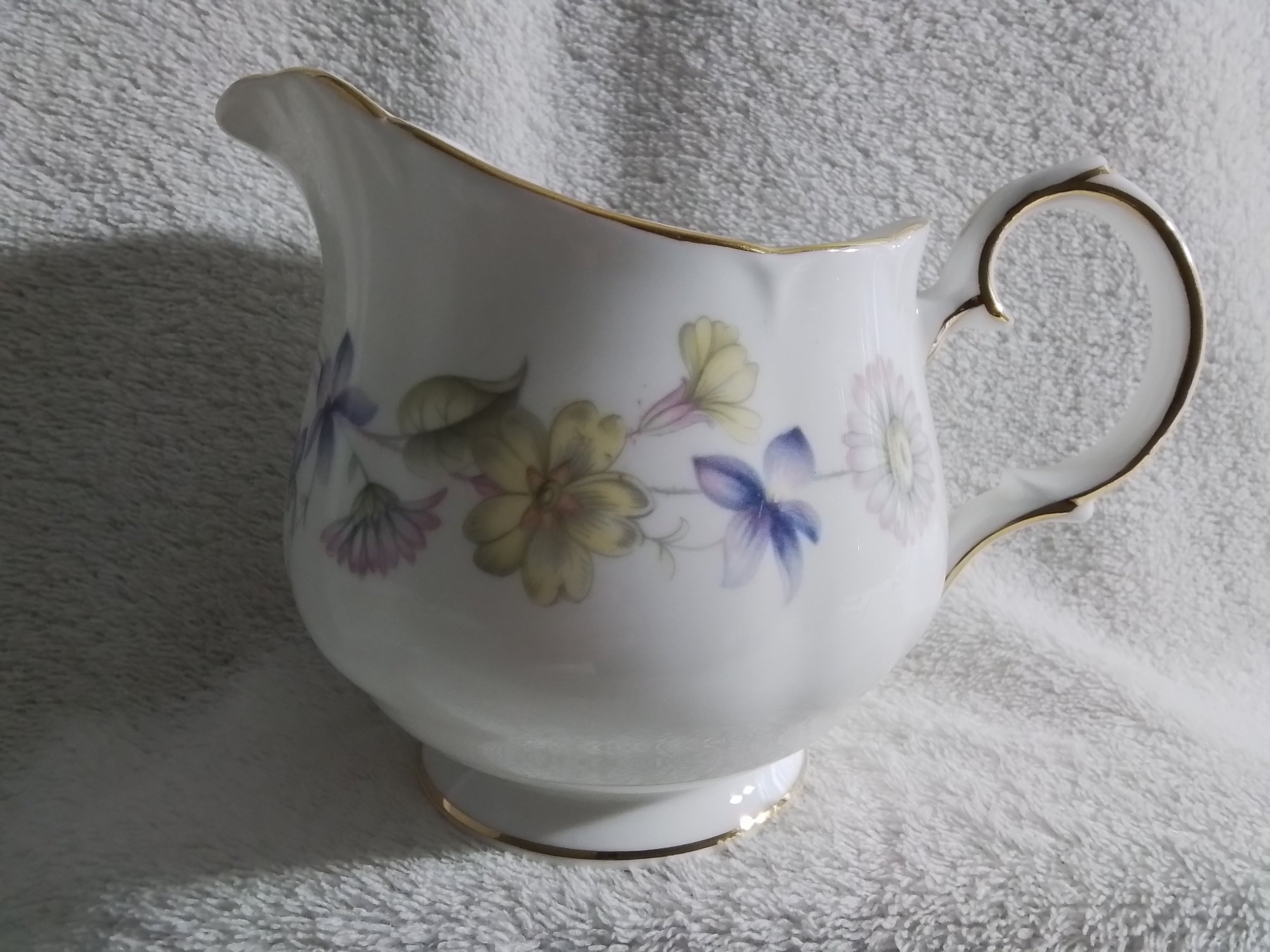 A porcelain creamer with floral patterns and gold accents on the rim, handle, and foot, placed on a soft fuzzy white surface.