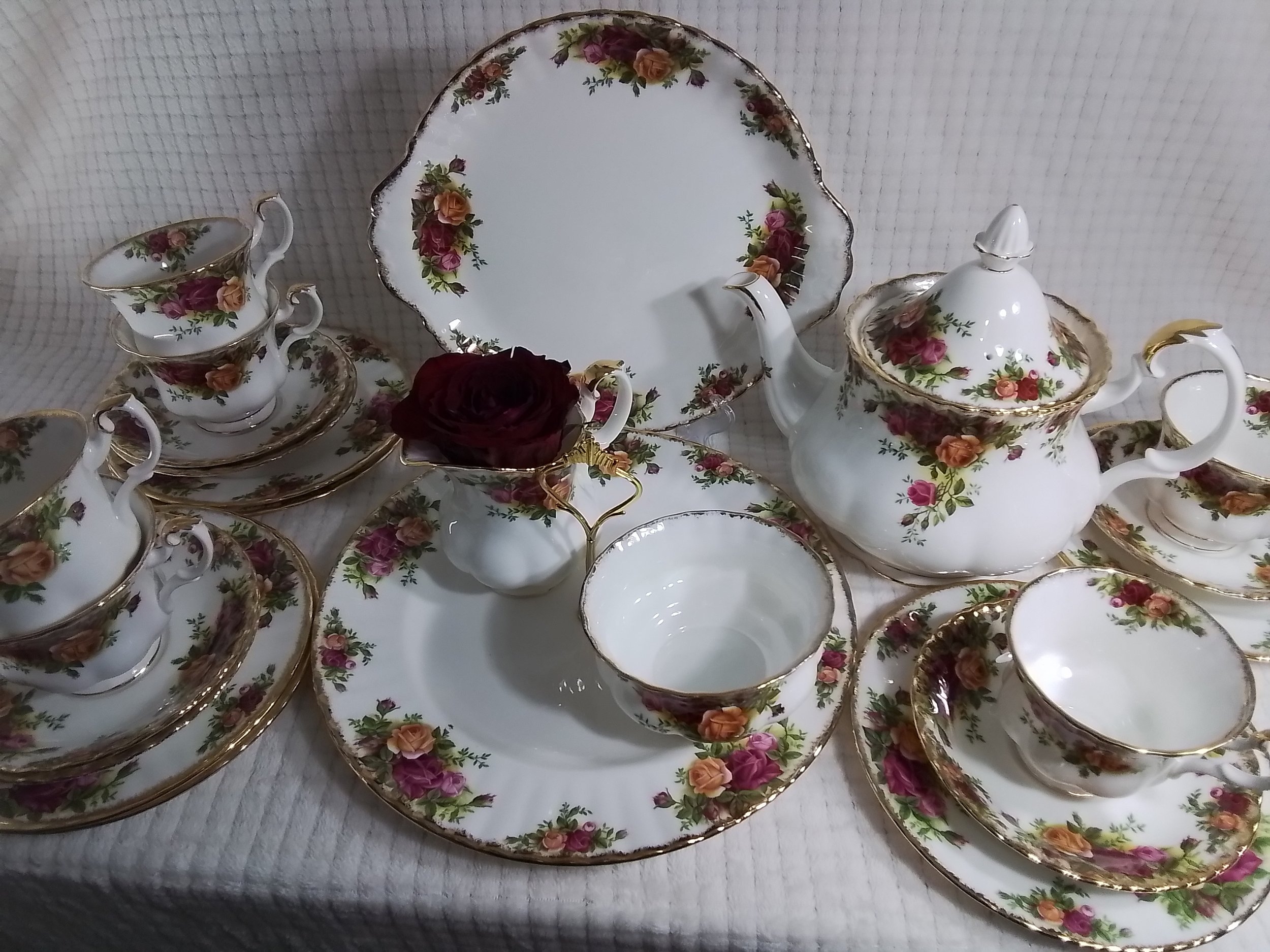 A vintage floral tea set on a white tablecloth, including a teapot, cups, saucers, a large plate, a small bowl, and a small creamer, with a dark red rose in a cup.