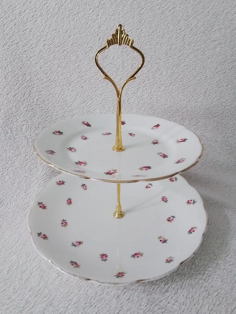 A two-tiered porcelain serving stand with a gold-colored metal handle and supports, decorated with small pink roses on both plates, against a textured light gray background.