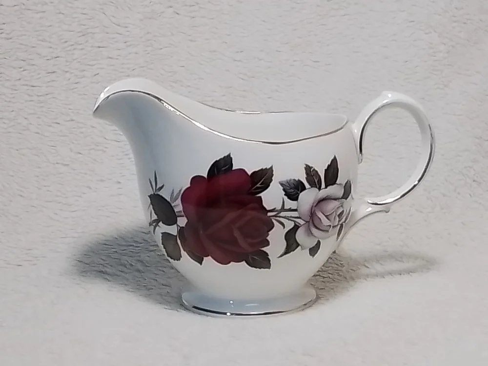 A white porcelain creamer with floral design featuring roses in shades of red and white, with green and black leaves.