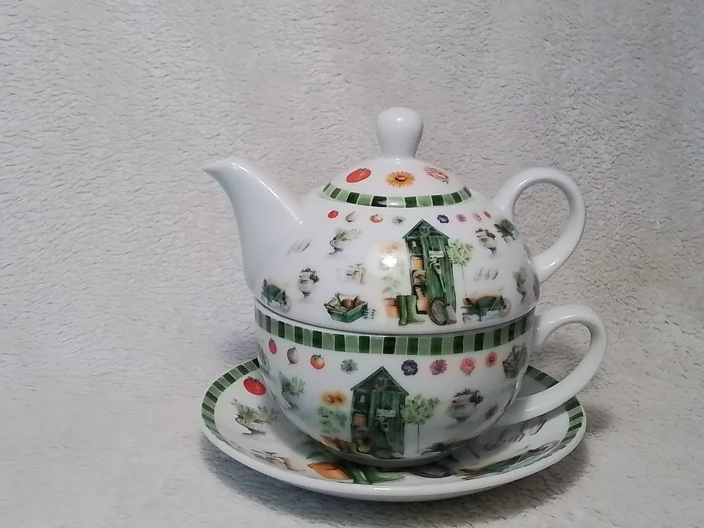 A white ceramic teapot with a lid, paired with a matching teacup, all on a matching saucer. The teapot has decorative images of garden sheds, flowers, and gardening tools, with green striped borders.
