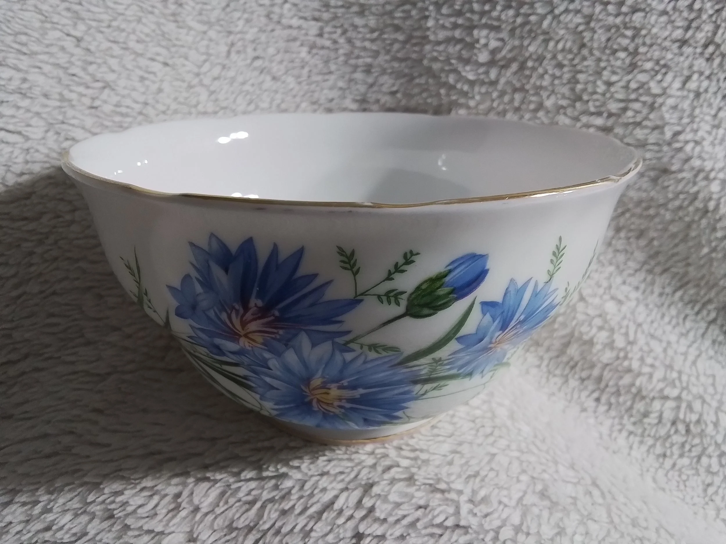 A white porcelain bowl with a gold rim, decorated with blue and purple flowers and green leaves, resting on a textured gray fabric background.