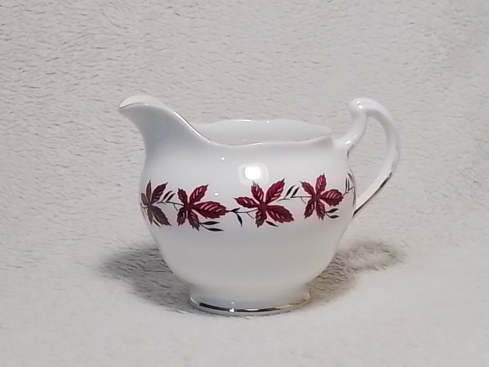 White ceramic creamer with red and black leaf design on a soft, fluffy surface.