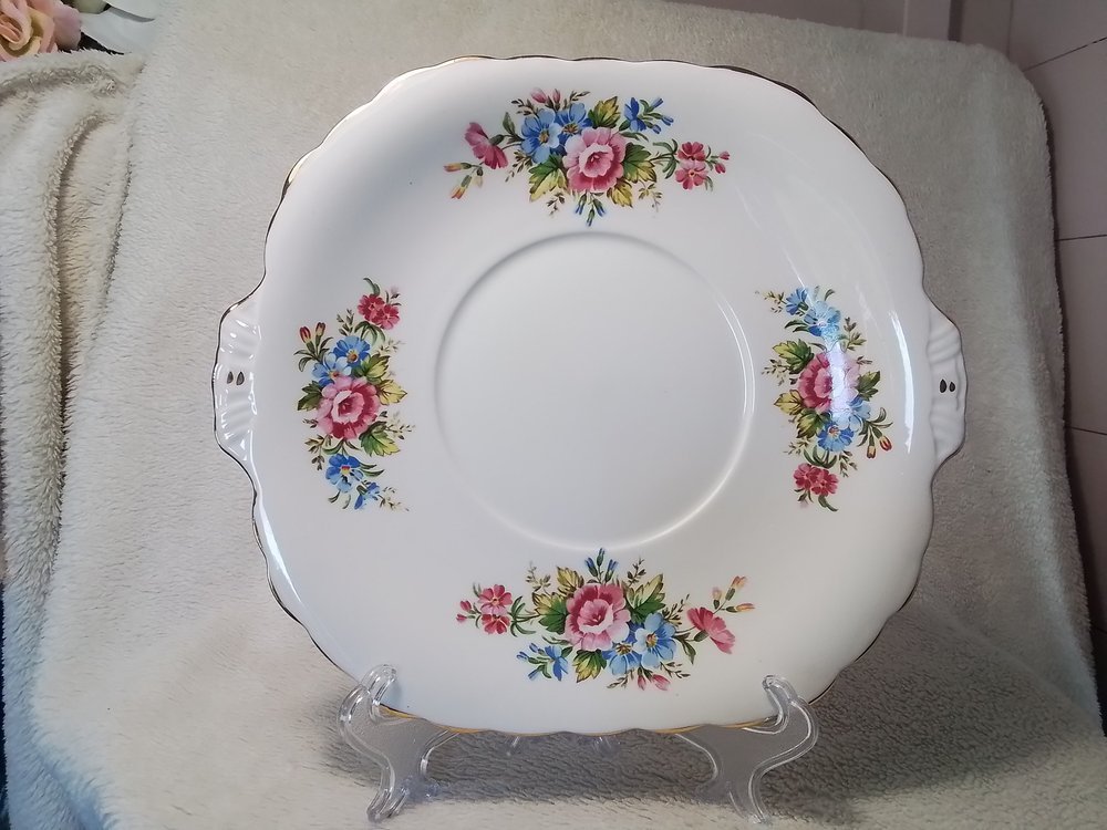 A decorative ceramic plate with a floral pattern of pink and blue flowers and green leaves, displayed on a clear stand on a soft, off-white surface.