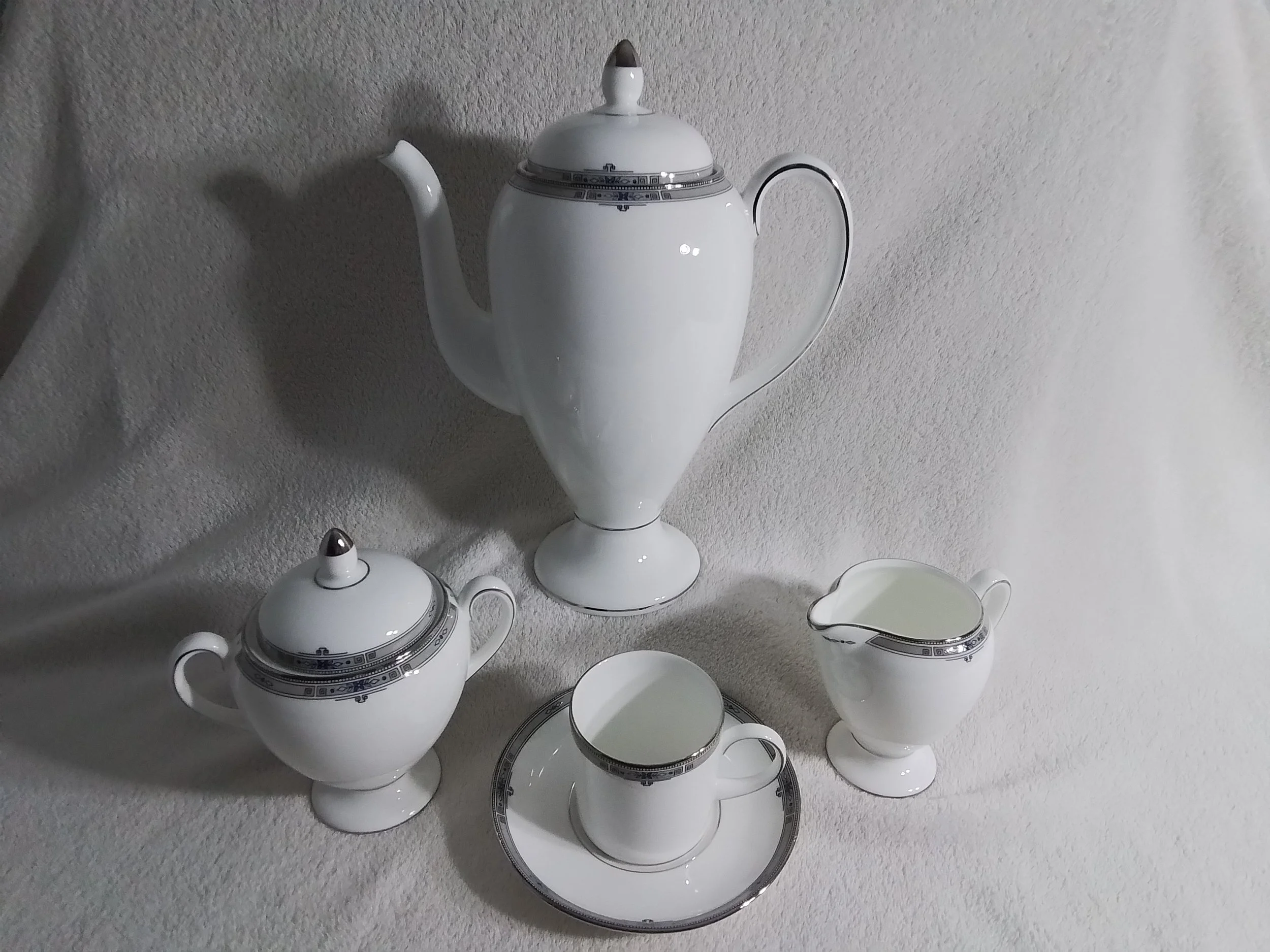 White porcelain tea set with black and silver decorative accents, including a teapot, sugar bowl with lid, creamer, a cup on a saucer, all arranged on a light-colored textured surface.