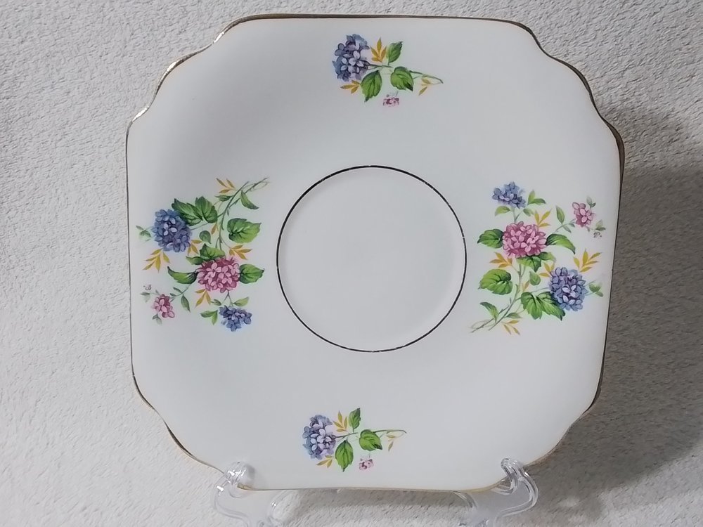 A white square ceramic plate with scalloped edges and a gold trim, decorated with purple and pink floral patterns.