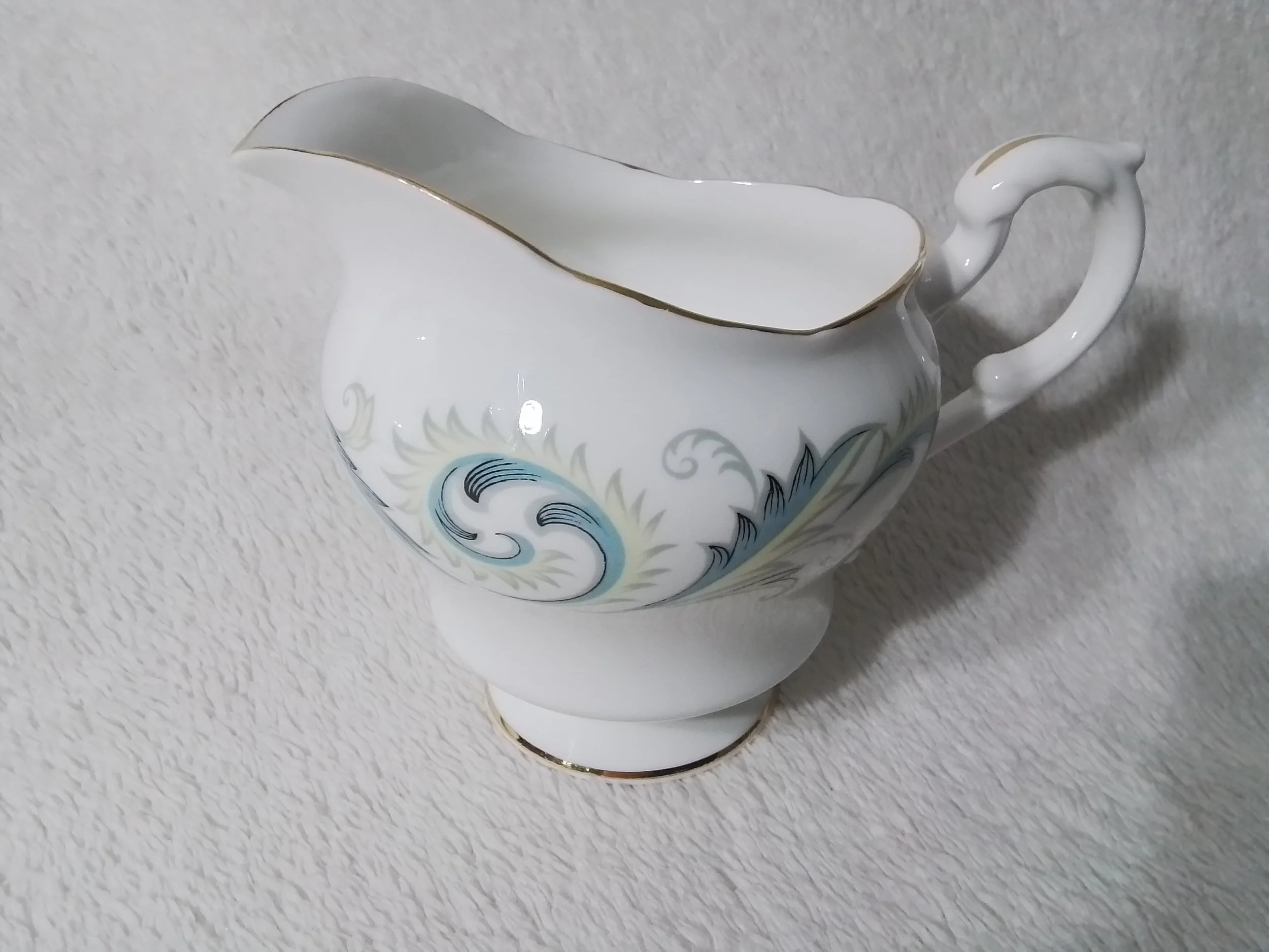 White porcelain gravy boat with gold trim and decorative blue and gray swirling pattern, positioned on a light textured surface.